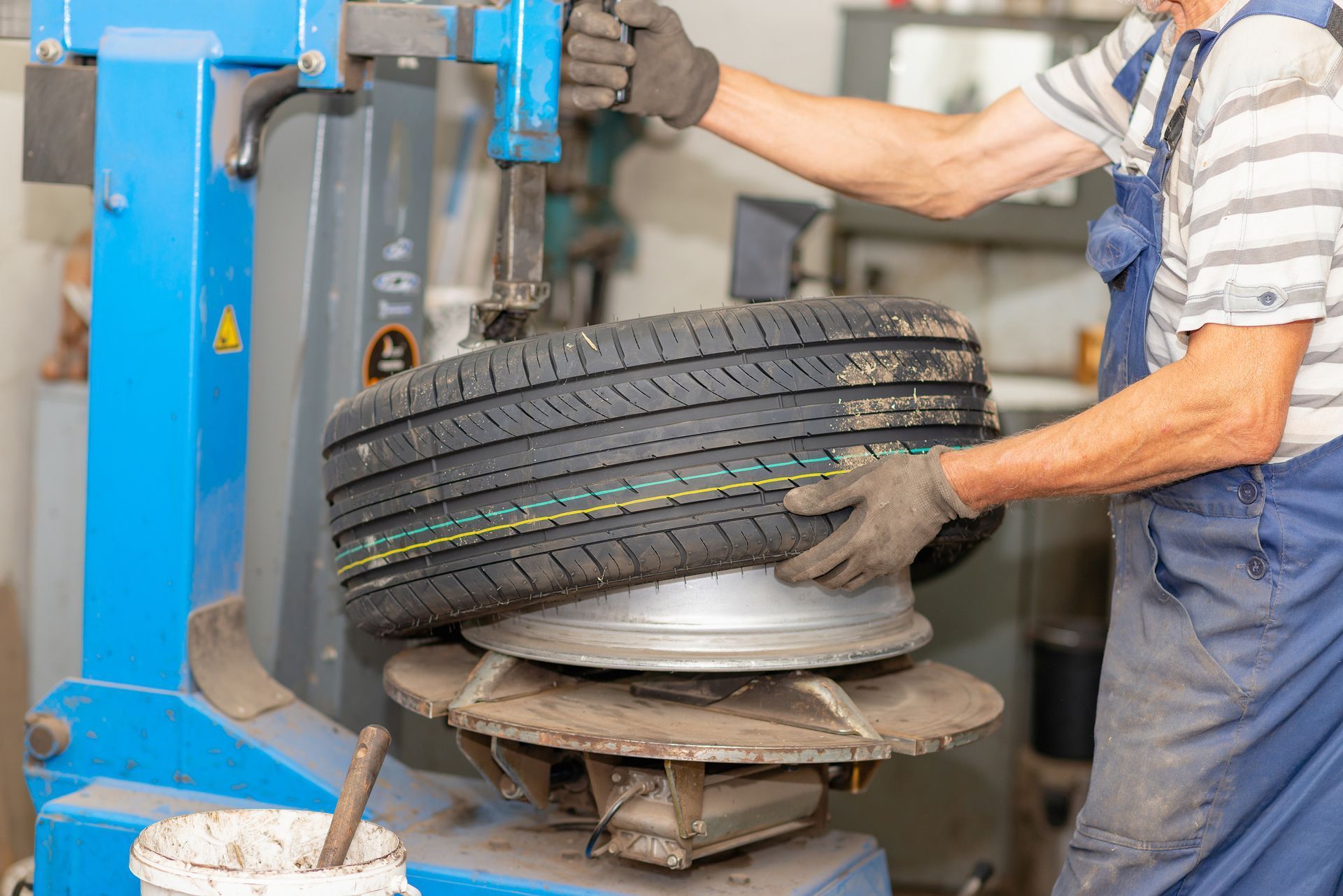 A technician in blue overalls uses a blue tire changing machine to mount a new tire onto a wheel rim in a workshop.