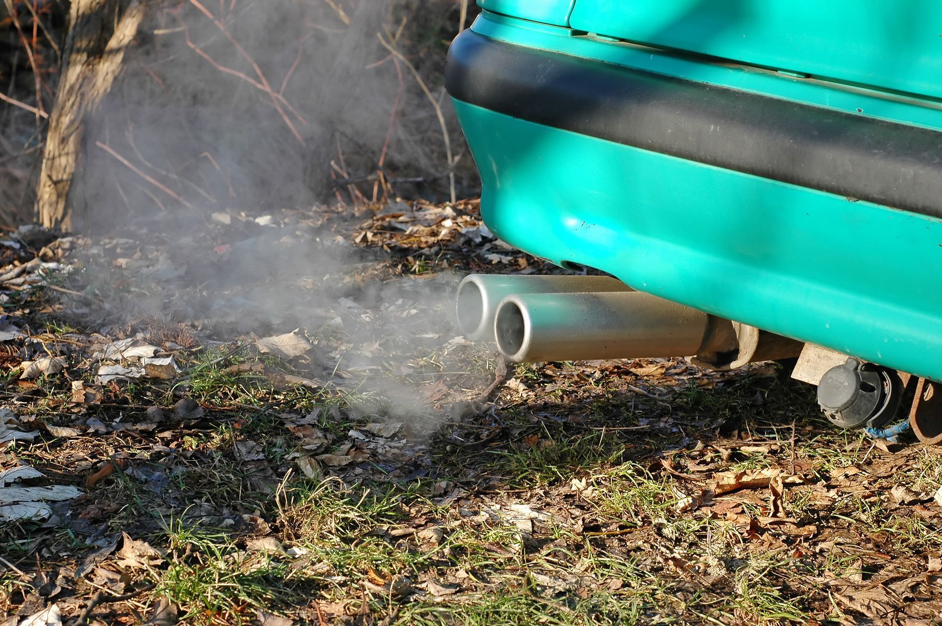 A teal car’s exhaust pipes emit a cloud of white smoke over dry grass and leaves.