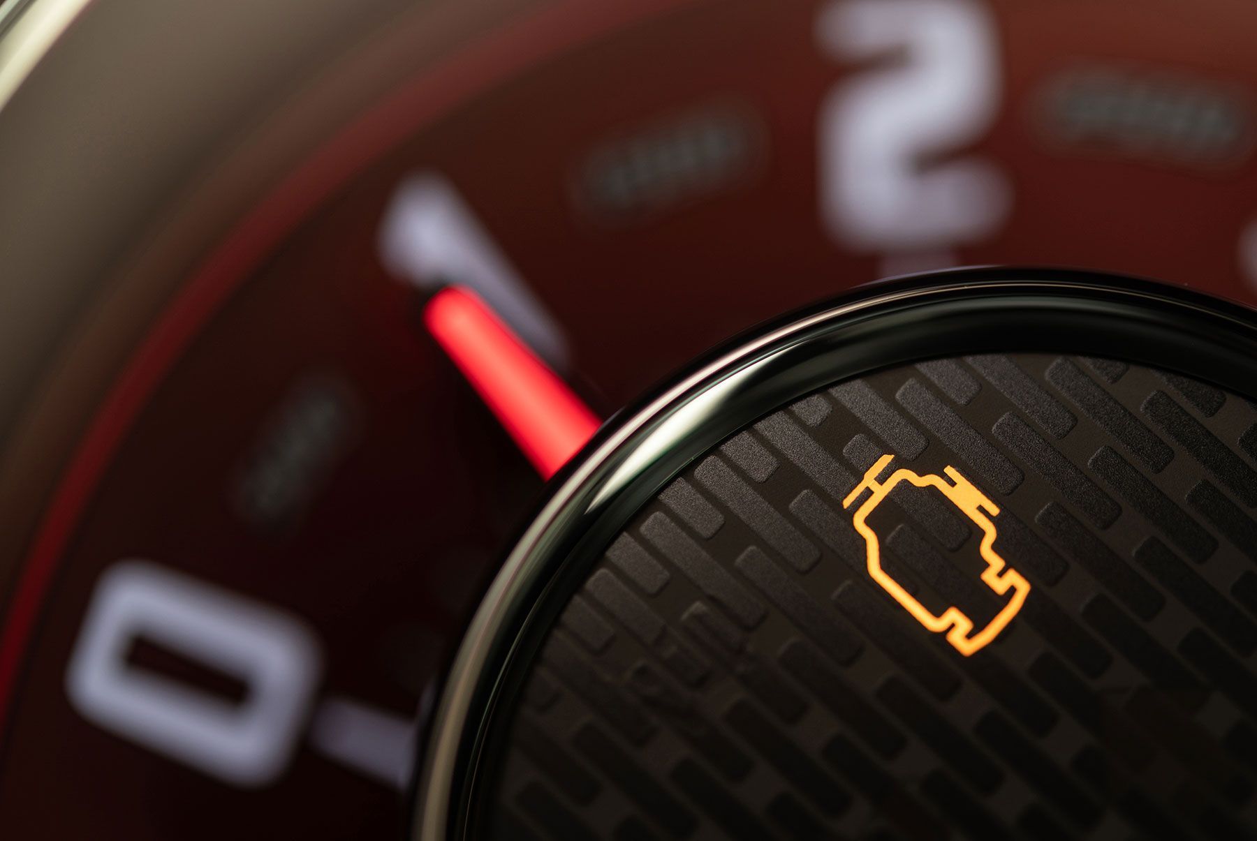 A glowing amber check engine light on a dark, textured circular display in front of a blurred car tachometer.