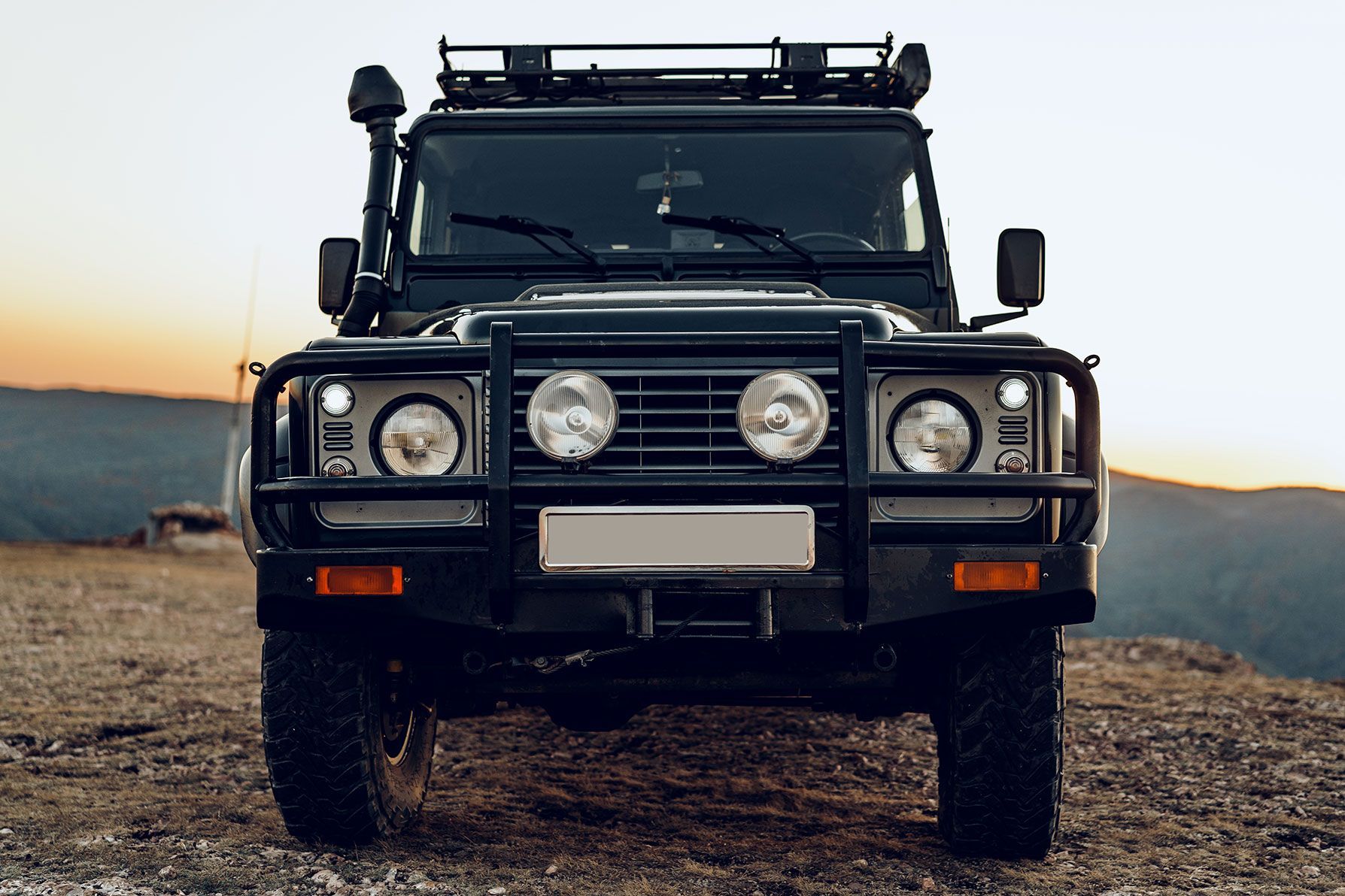 A black off-road vehicle with a snorkel and roof rack parked on a rocky terrain during sunset.