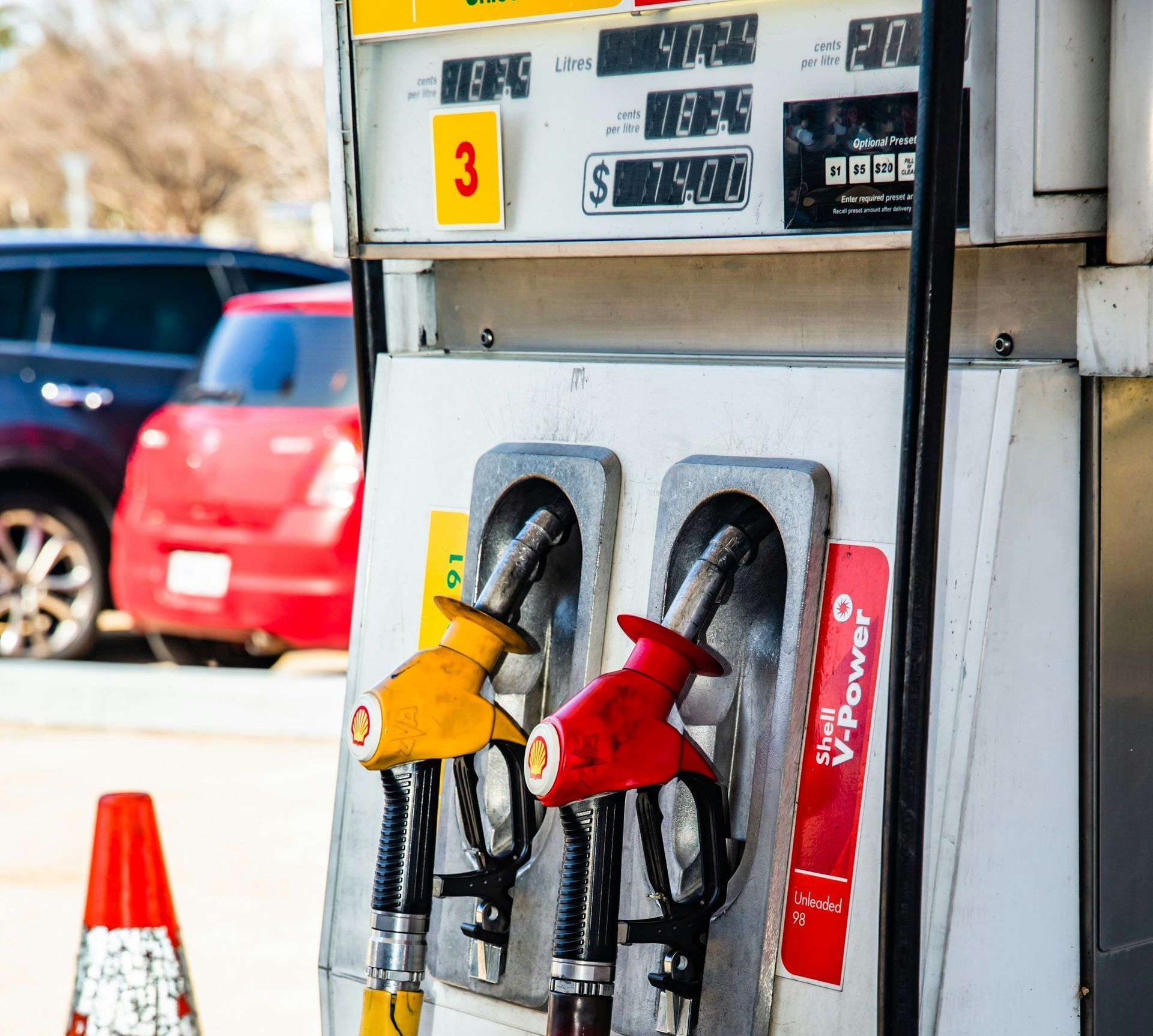 A gas pump with a yellow and a red nozzle in the foreground, with a partial view of cars in the background.