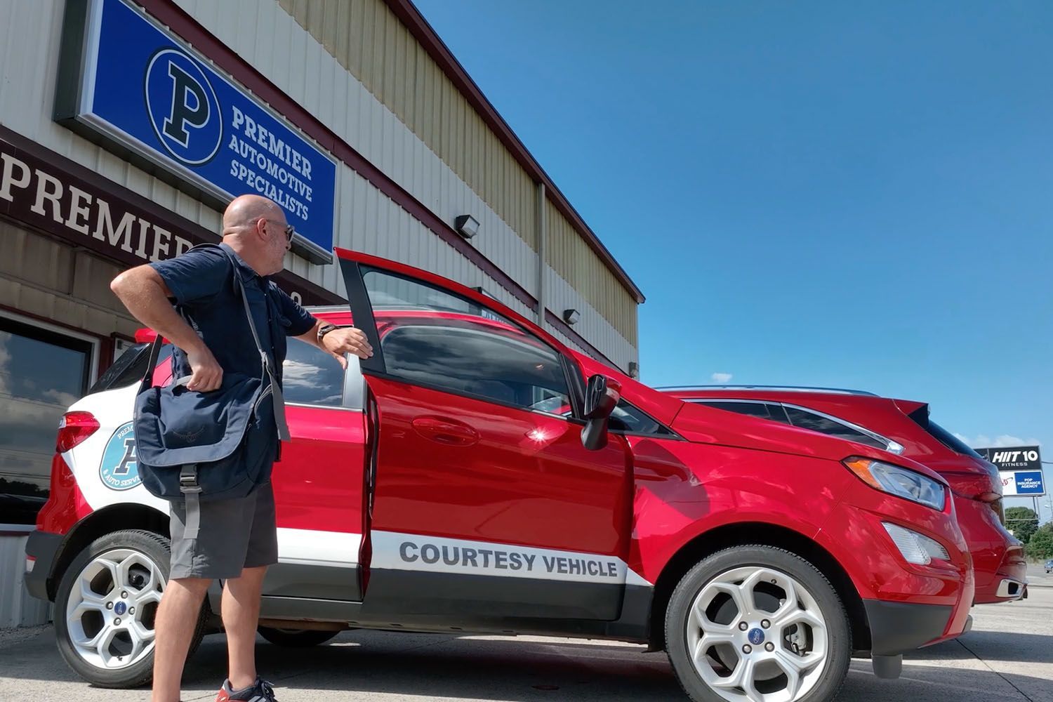 Man standing by a red car with open door outside a storefront on a sunny day