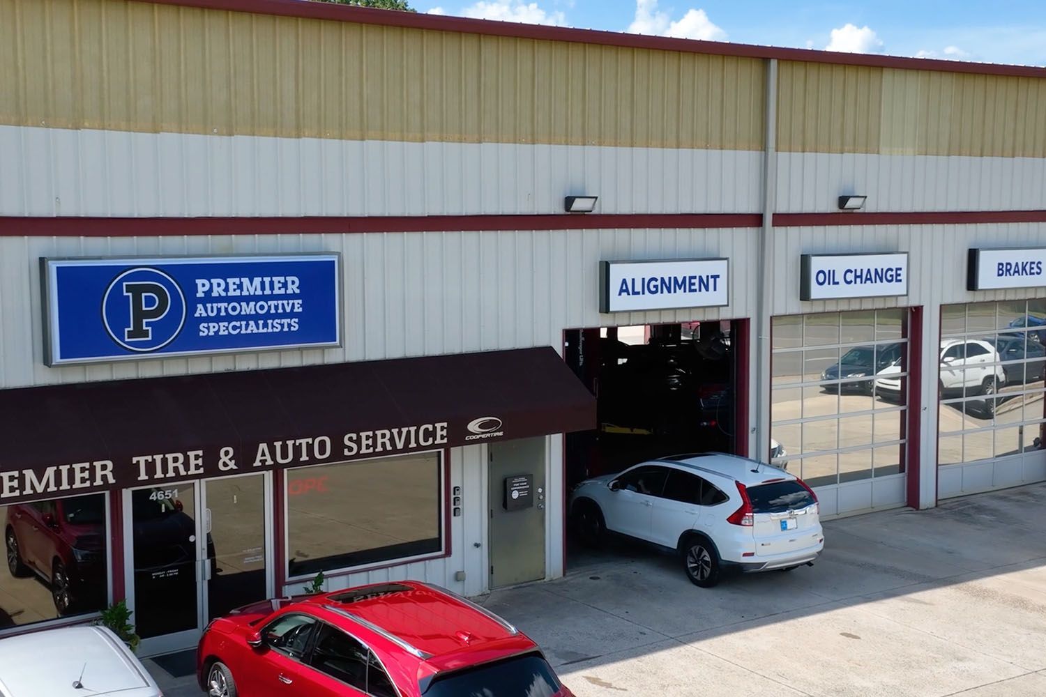 Auto repair shop exterior with service bays and parked cars, including a red car and a white SUV