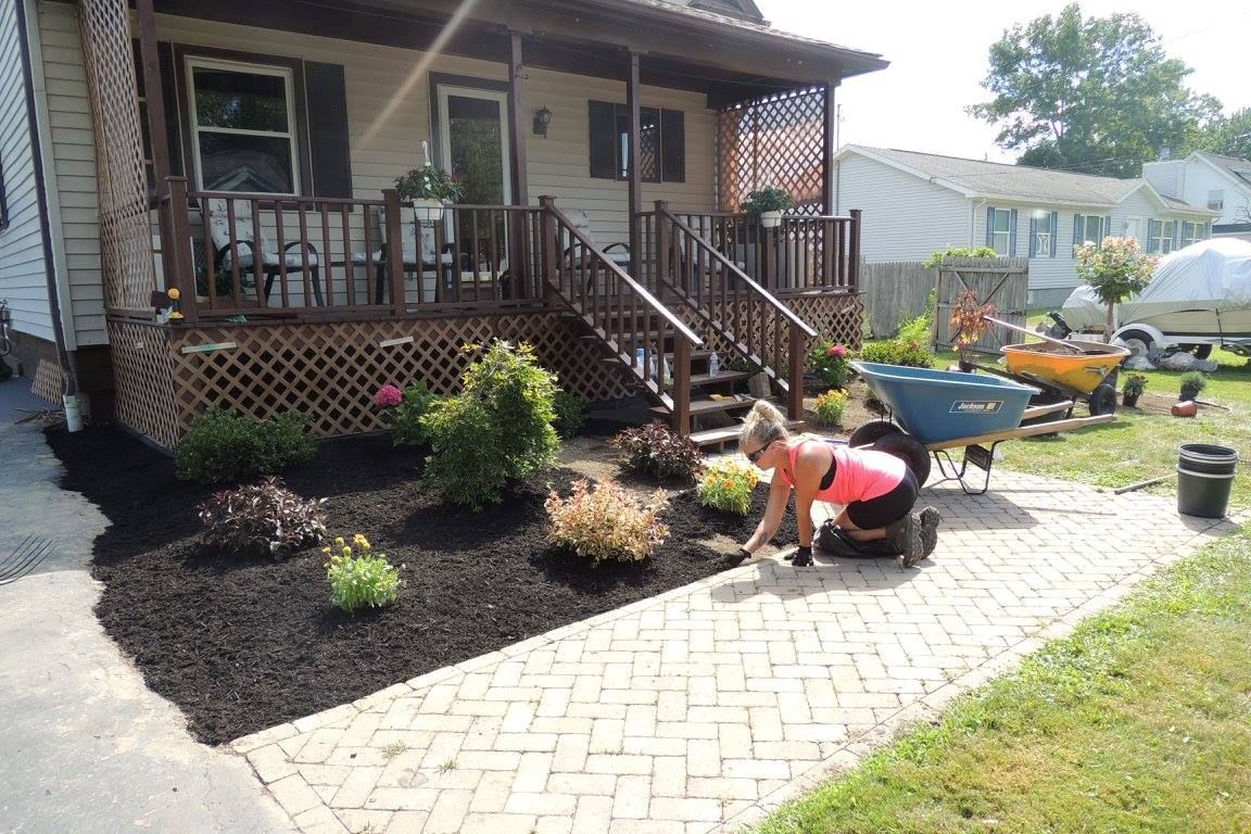 Person planting flowers in a front yard beside a brick walkway and porch