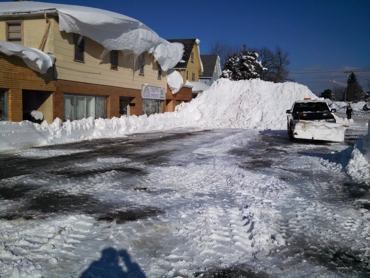 Snow-covered residential street with a yellow house and a car near a large plowed snowbank.