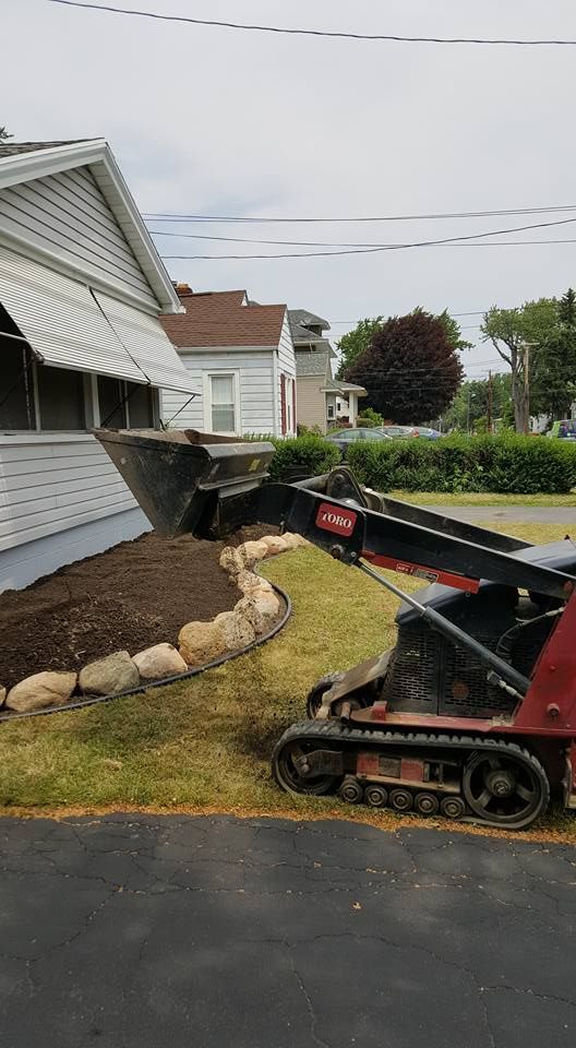 Mini excavator digging beside a house, moving soil near a landscaped yard and driveway.