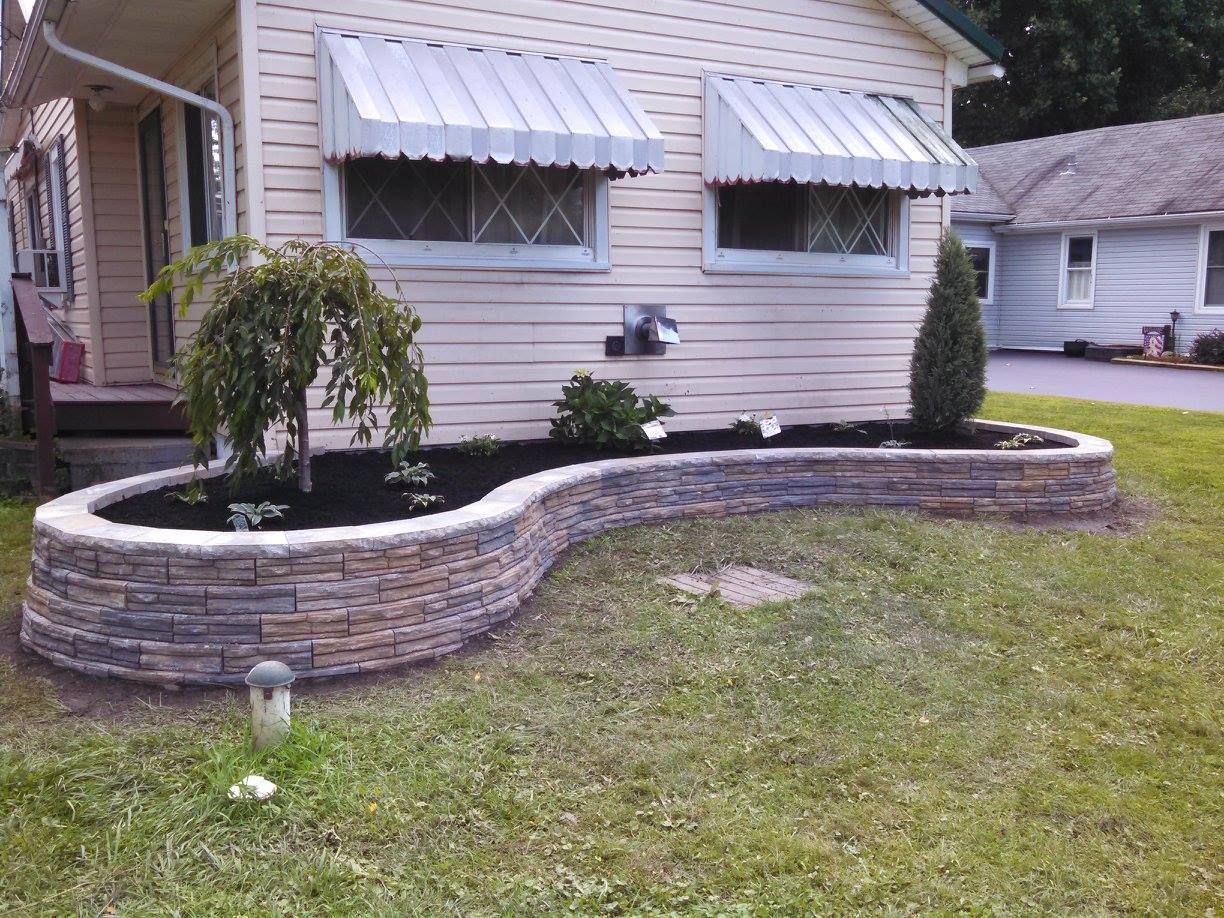 Front yard with a small tree in a raised stone planter beside a white house with awnings
