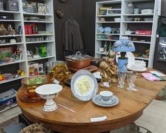 Antique decor on a round table in a thrift shop, with decorative plates, glassware, and a lamp.