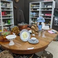 Vintage round table display with plates, teacups, lamp, and shelves of home decor in a shop