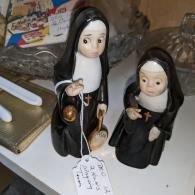Two small nun figurines in black habits standing on a white surface, one holding a rosary and lamp.