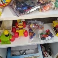 Toy shelf with colorful toy vehicles, figures, and small parts arranged on white shelves.