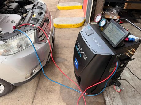 Car battery jump starter connected by red and blue cables to a car in a driveway, with a diagnostic display.