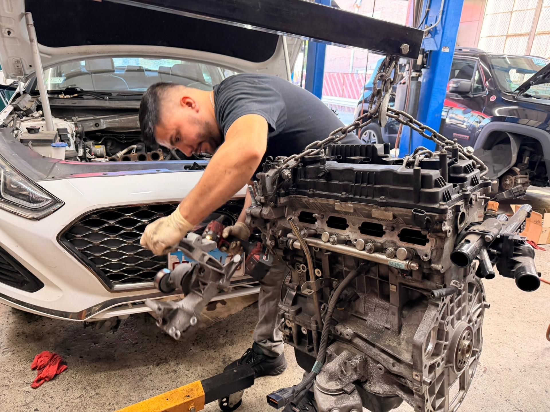 Mechanic working under a raised car hood beside a detached engine in an auto repair shop