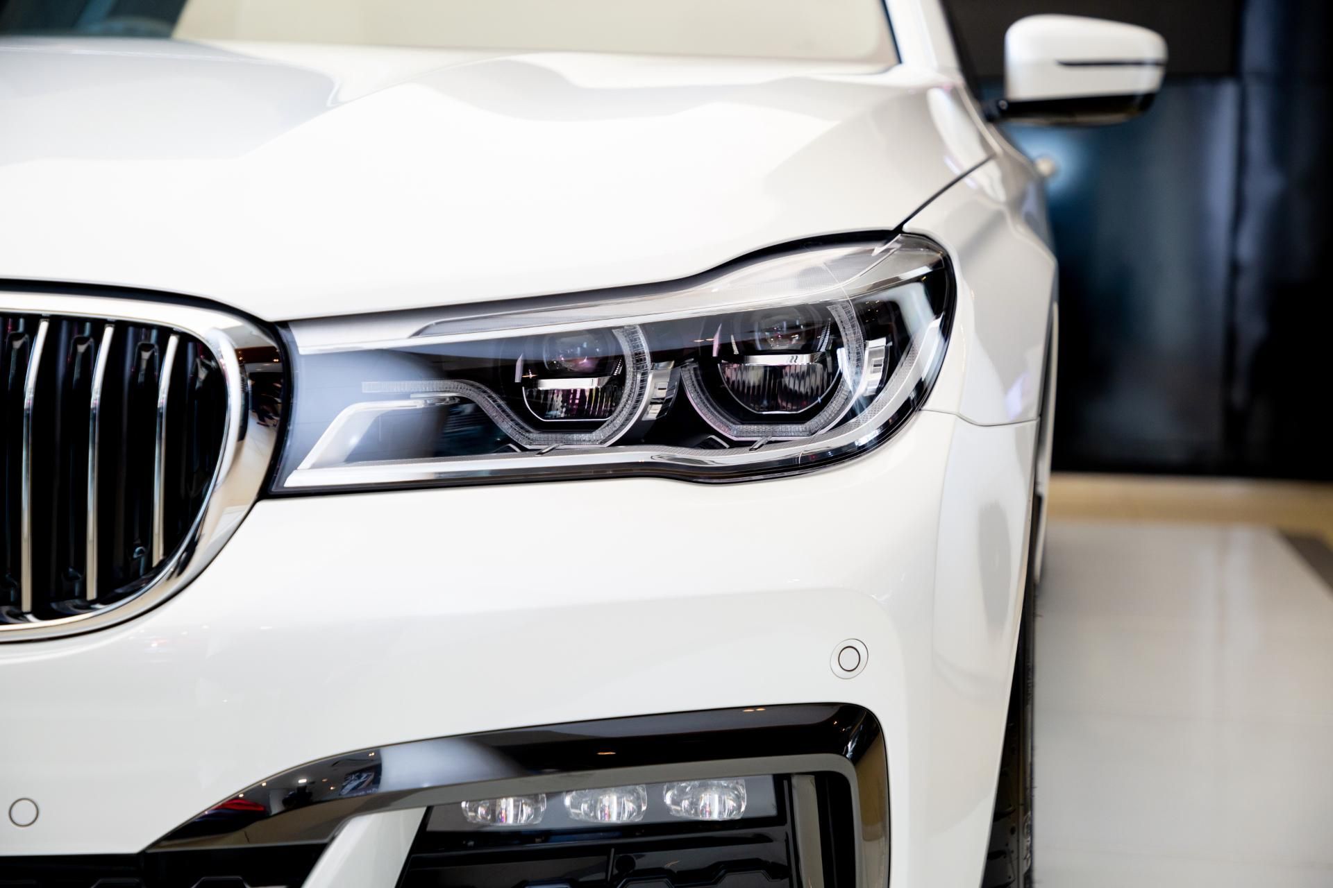 Front-left close-up of a white BMW sedan, showing the headlight and grille in a showroom
