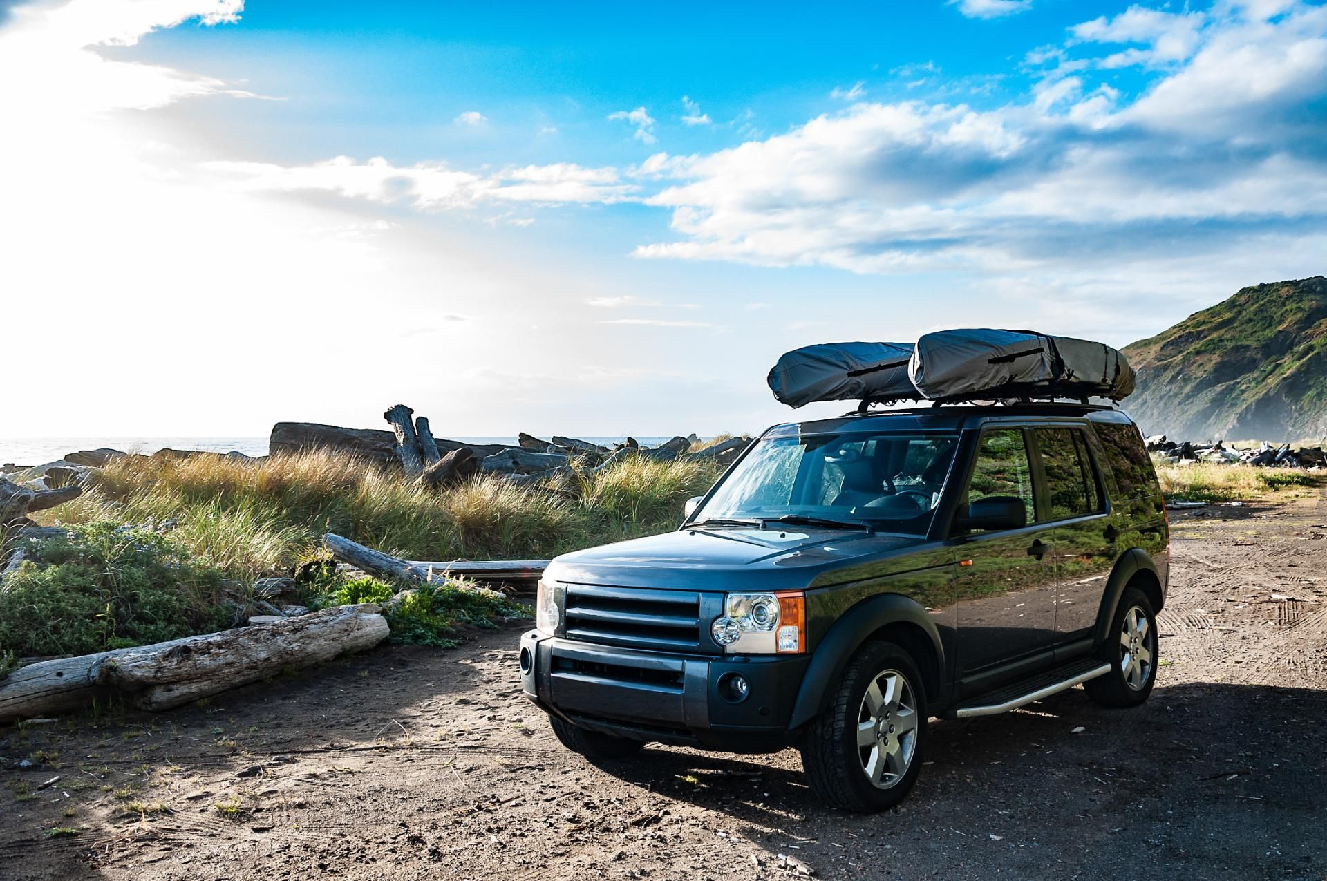 Blue SUV with roof cargo parked on a coastal dirt road under a bright sky