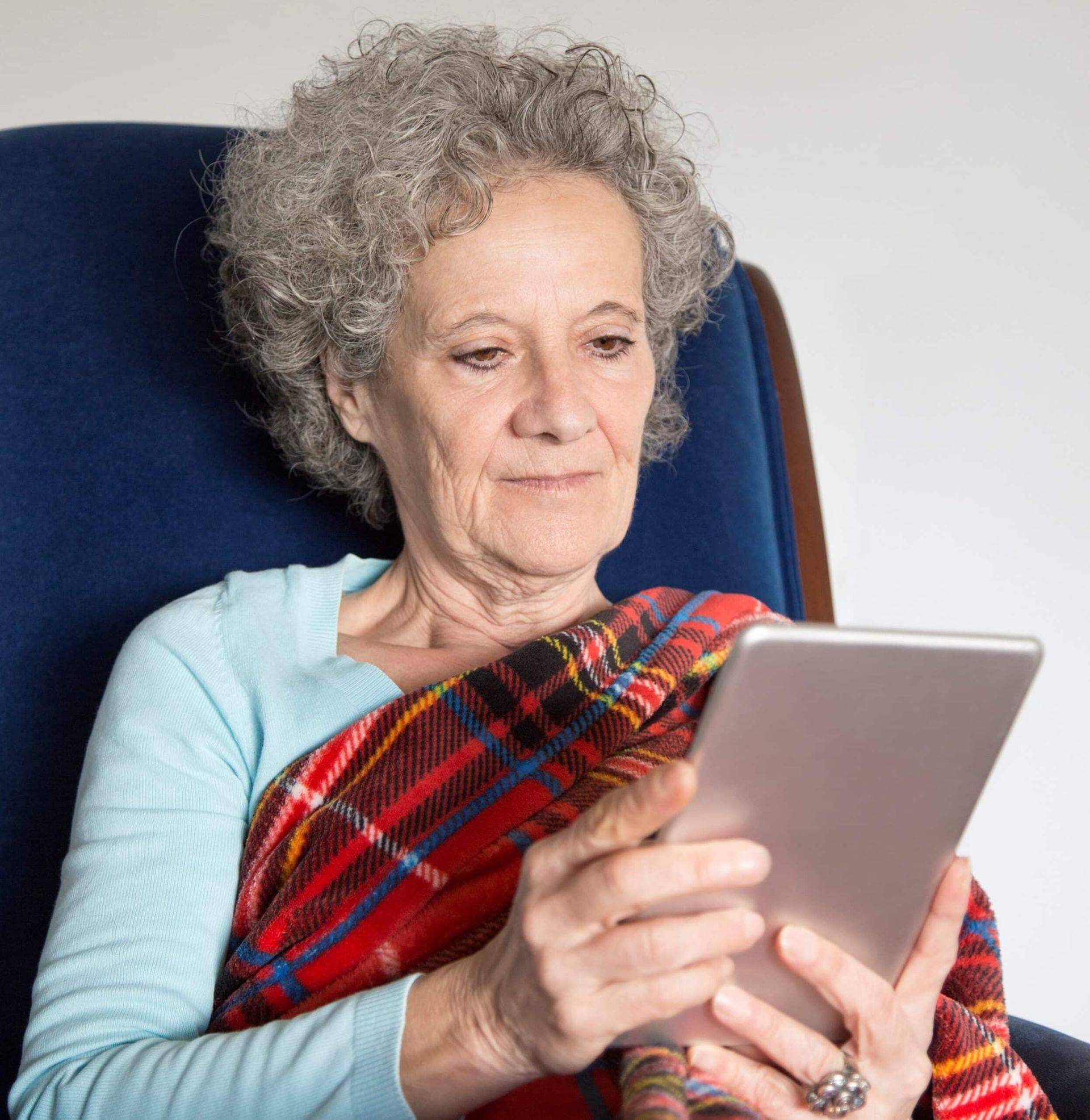 Elderly woman with gray curly hair, wrapped in a blanket, sitting in a blue chair, using a tablet.