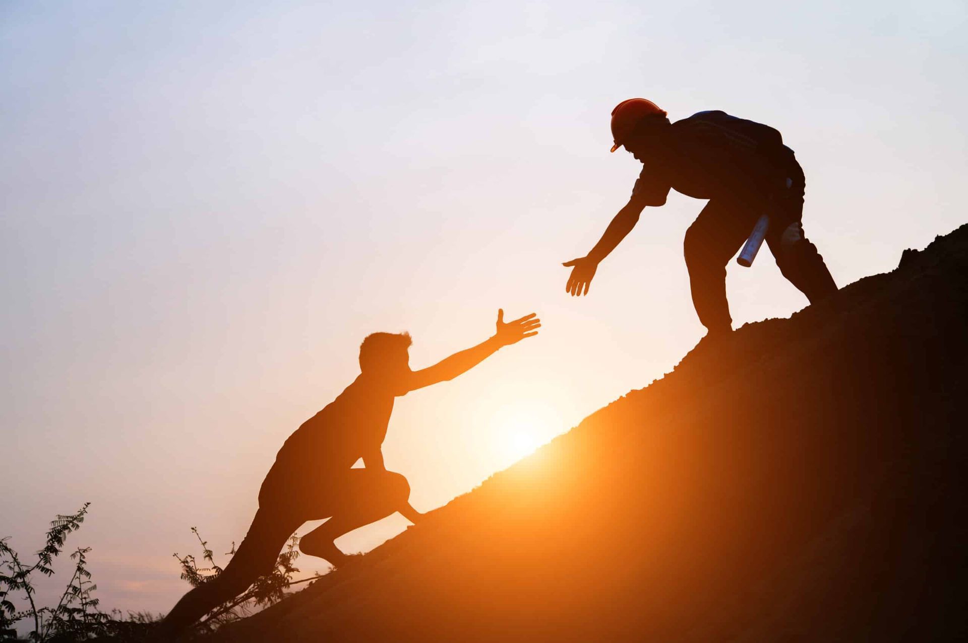 Silhouette of two people climbing a mountain, one reaching a hand to help the other. Sunset in the background.