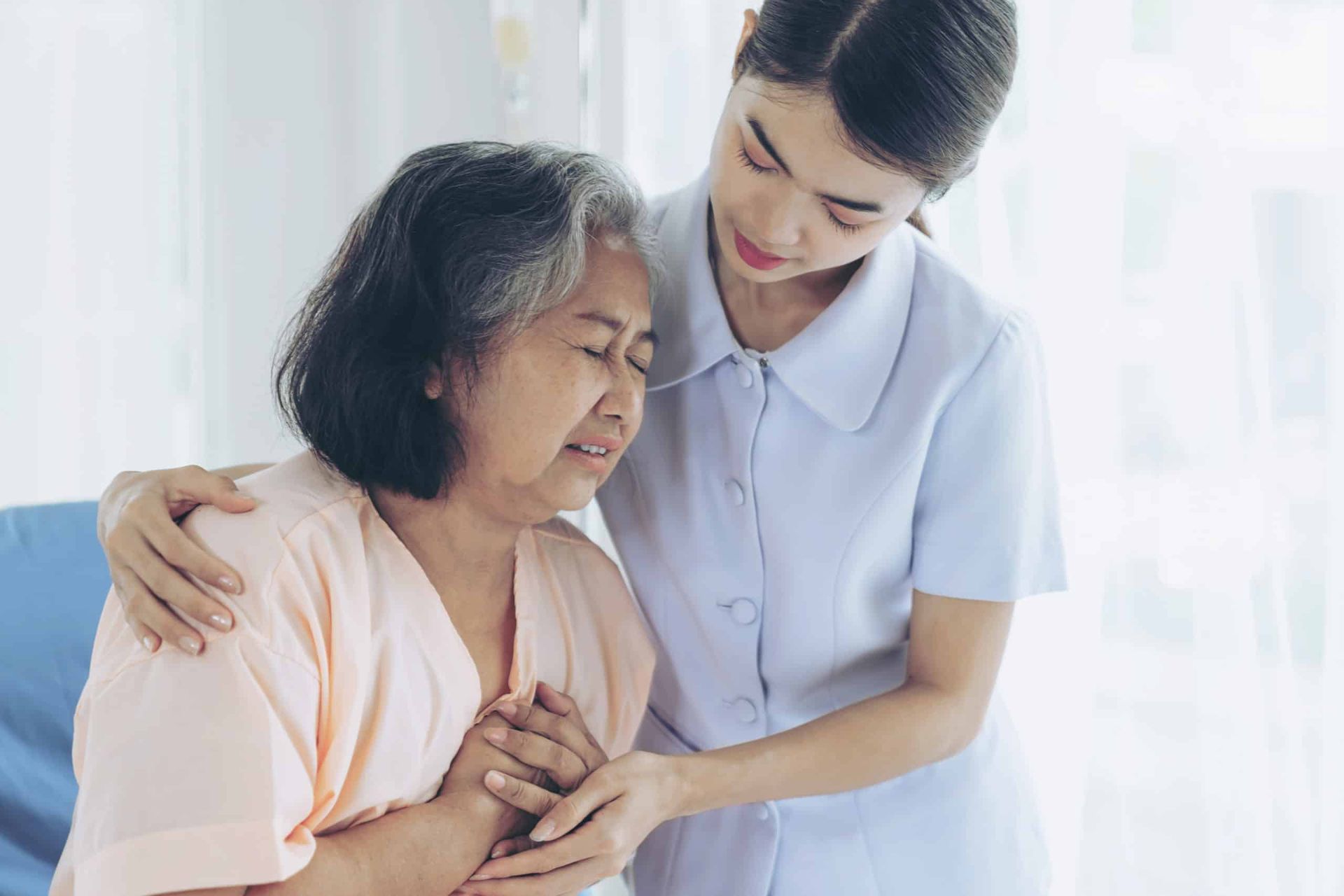Nurse comforting an elderly woman in a hospital room; nurse has arm around woman, holding her hands.