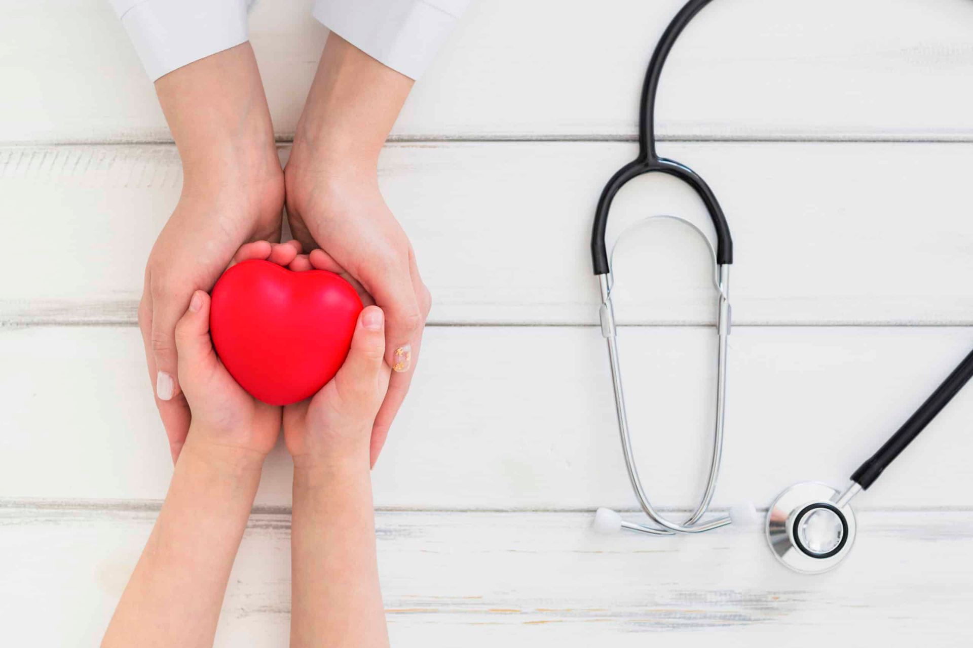 Hands holding a red heart with a stethoscope on a white wooden surface.