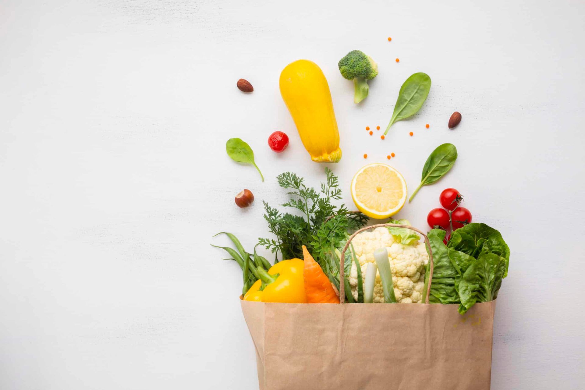 Brown paper bag overflowing with colorful fresh vegetables and fruit against a white background.