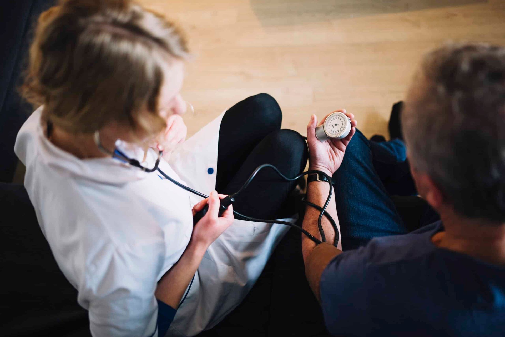 Nurse takes patient's blood pressure indoors on a couch. She wears white coat, he wears blue shirt.