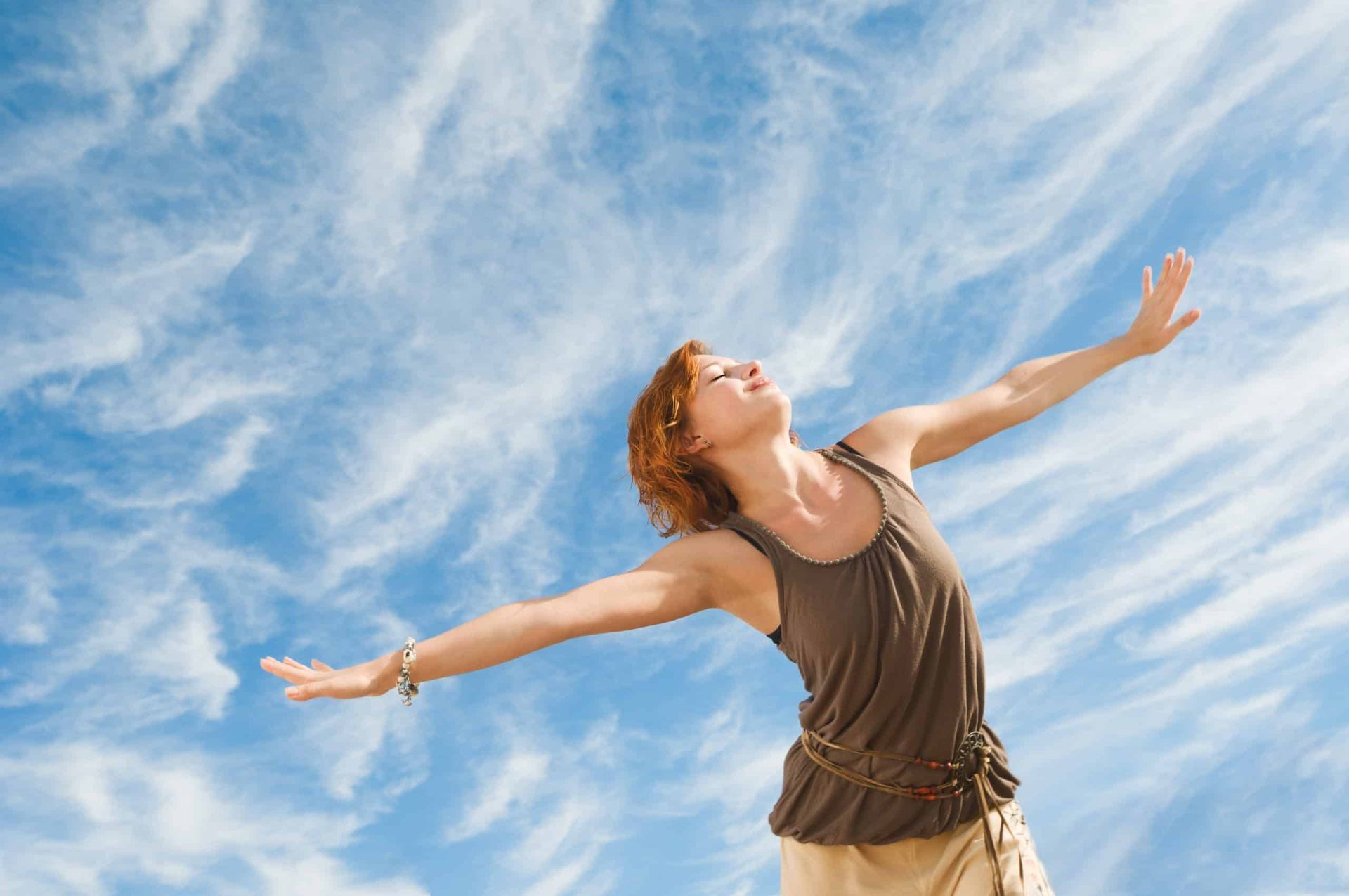Woman with arms outstretched, eyes closed, facing a blue sky with clouds.