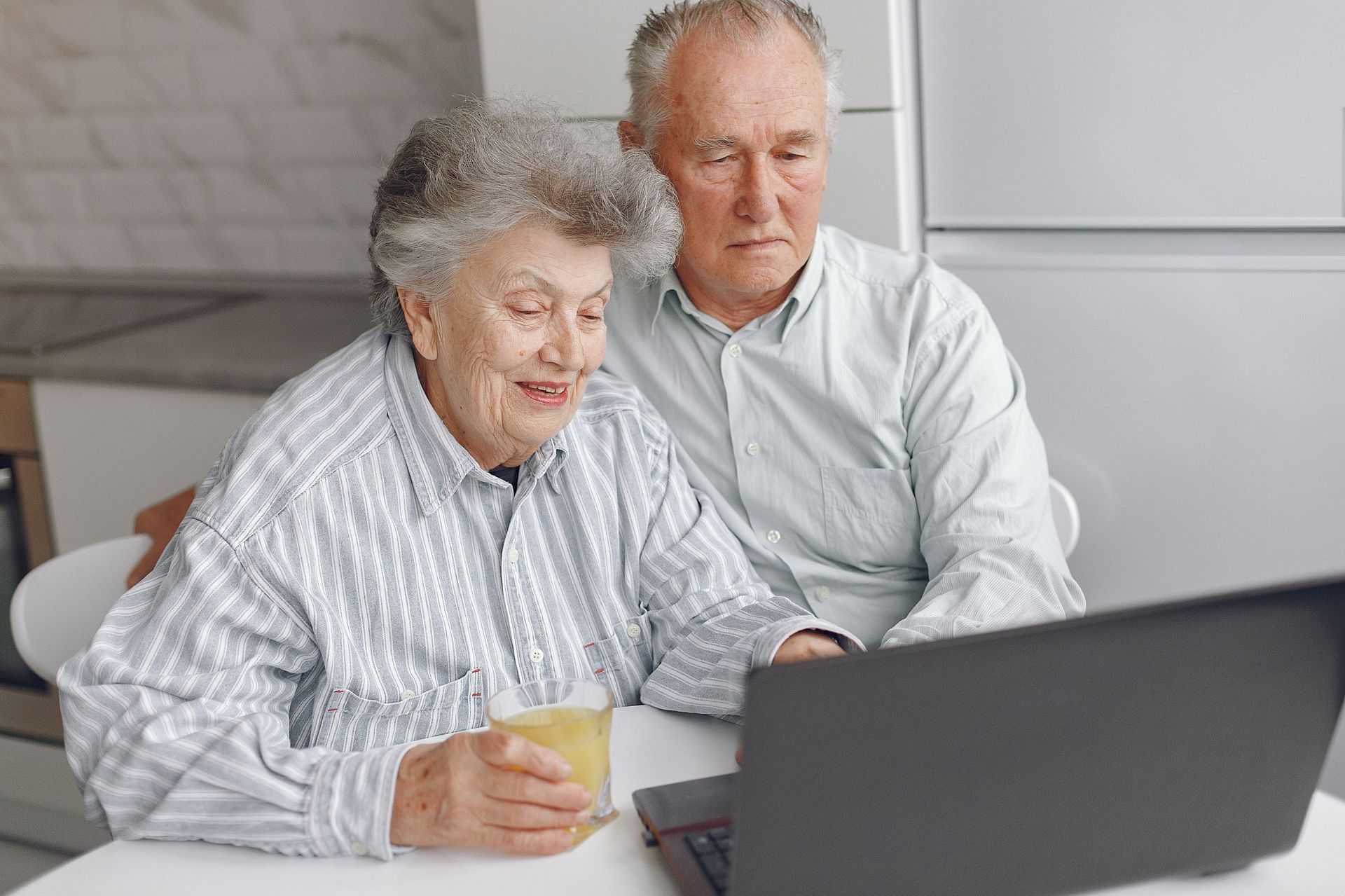 Elderly couple looking at a laptop together indoors. Woman holds a drink, both smiling.