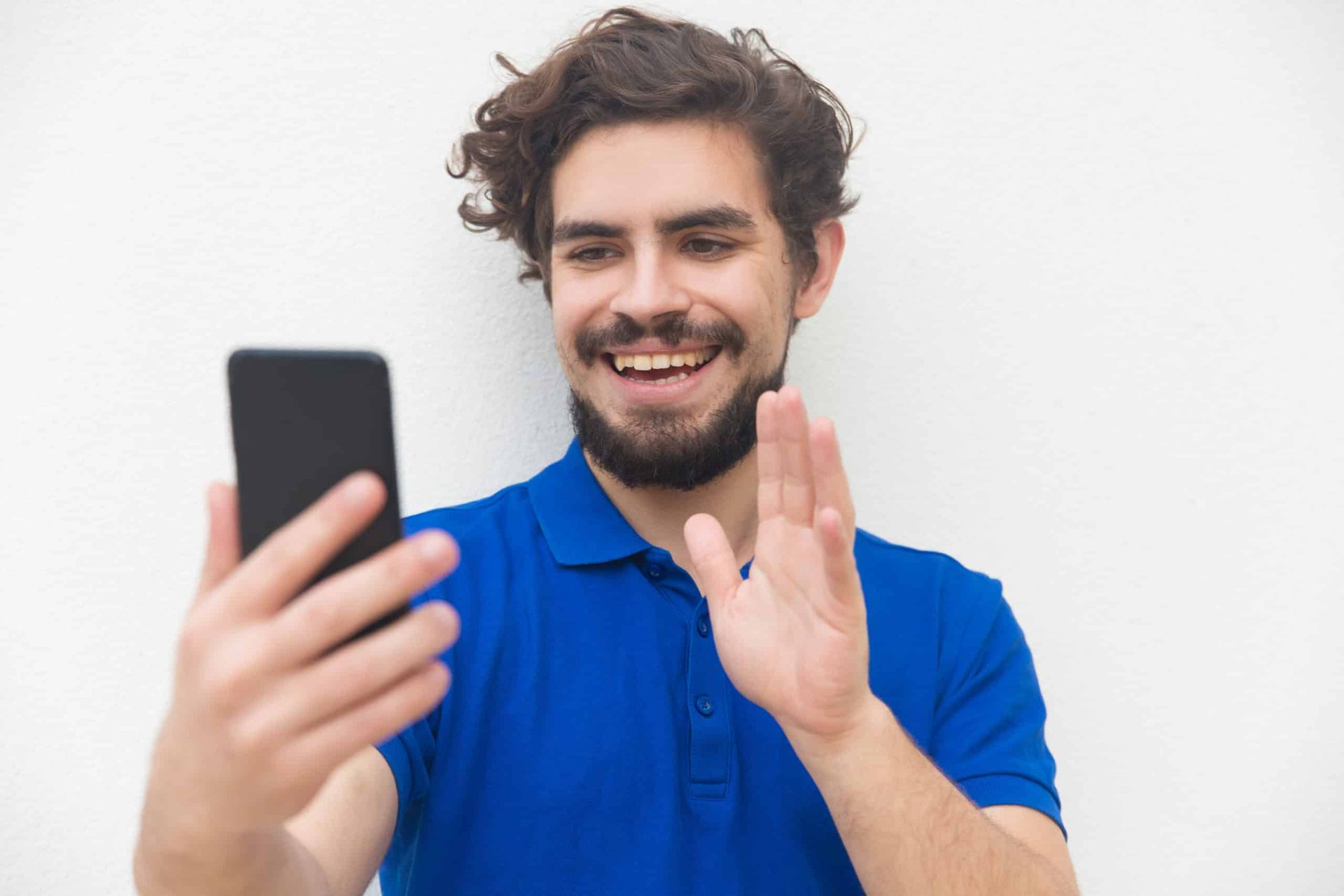 Man with beard waves while looking at a phone, smiling. Blue shirt against a white wall.
