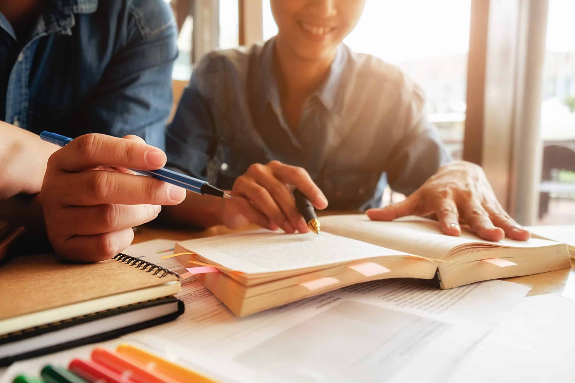 Two people studying at a desk, one pointing with a pen at an open book, another writing.
