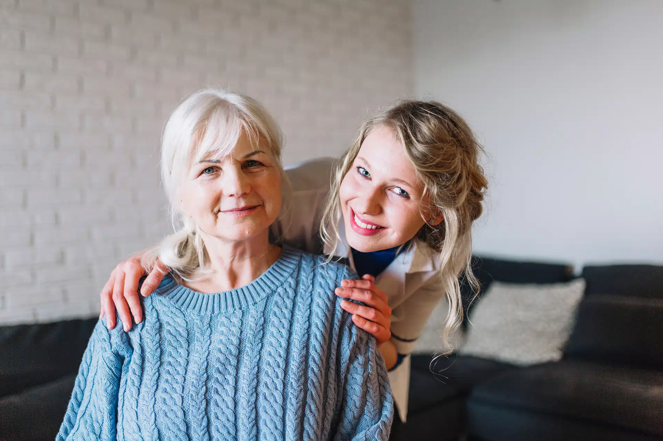 Woman with gray hair smiles, embraced by a younger woman with blonde hair, indoors.
