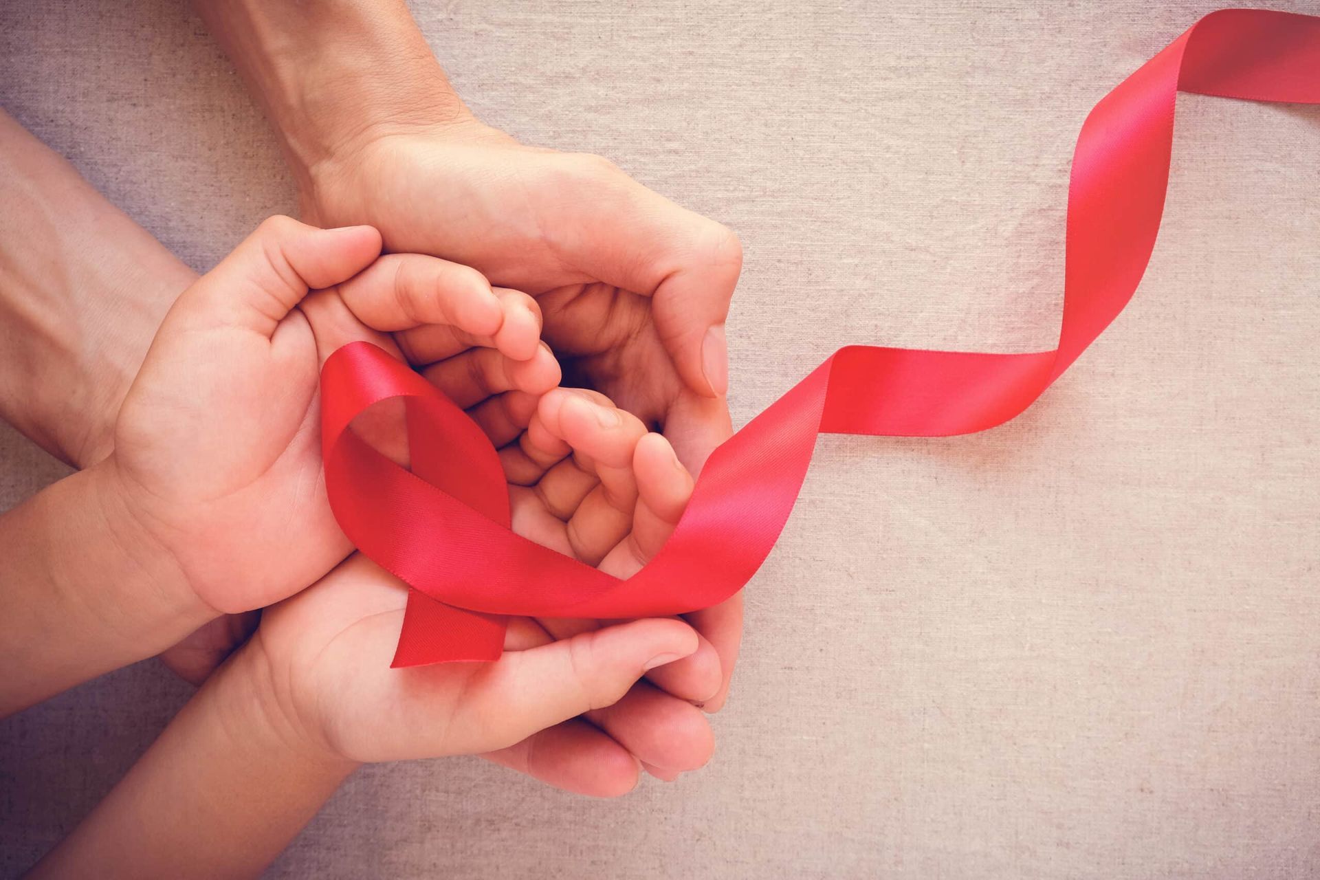 Hands holding a red ribbon, symbolizing AIDS awareness, on a cream-colored surface.
