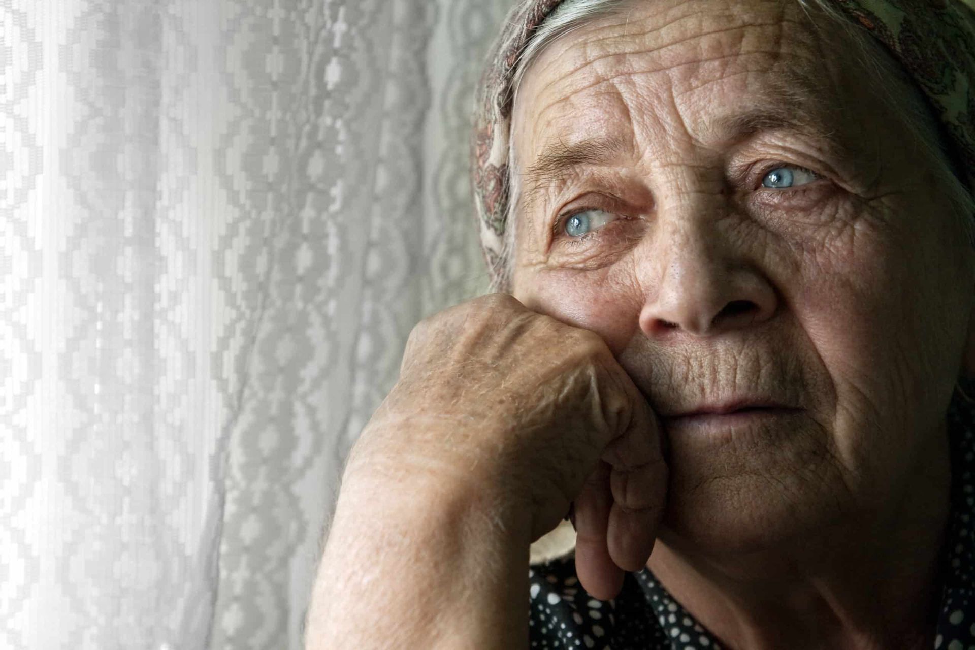 Elderly woman with thoughtful expression, resting chin on hand near a window.