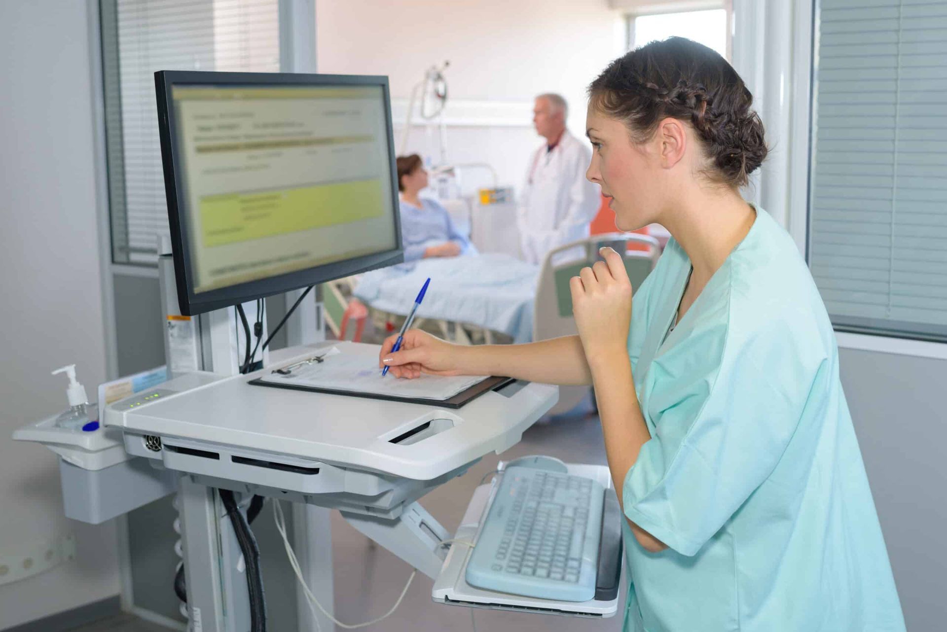 Nurse in scrubs, writing at computer on mobile workstation in hospital room, patient in bed.