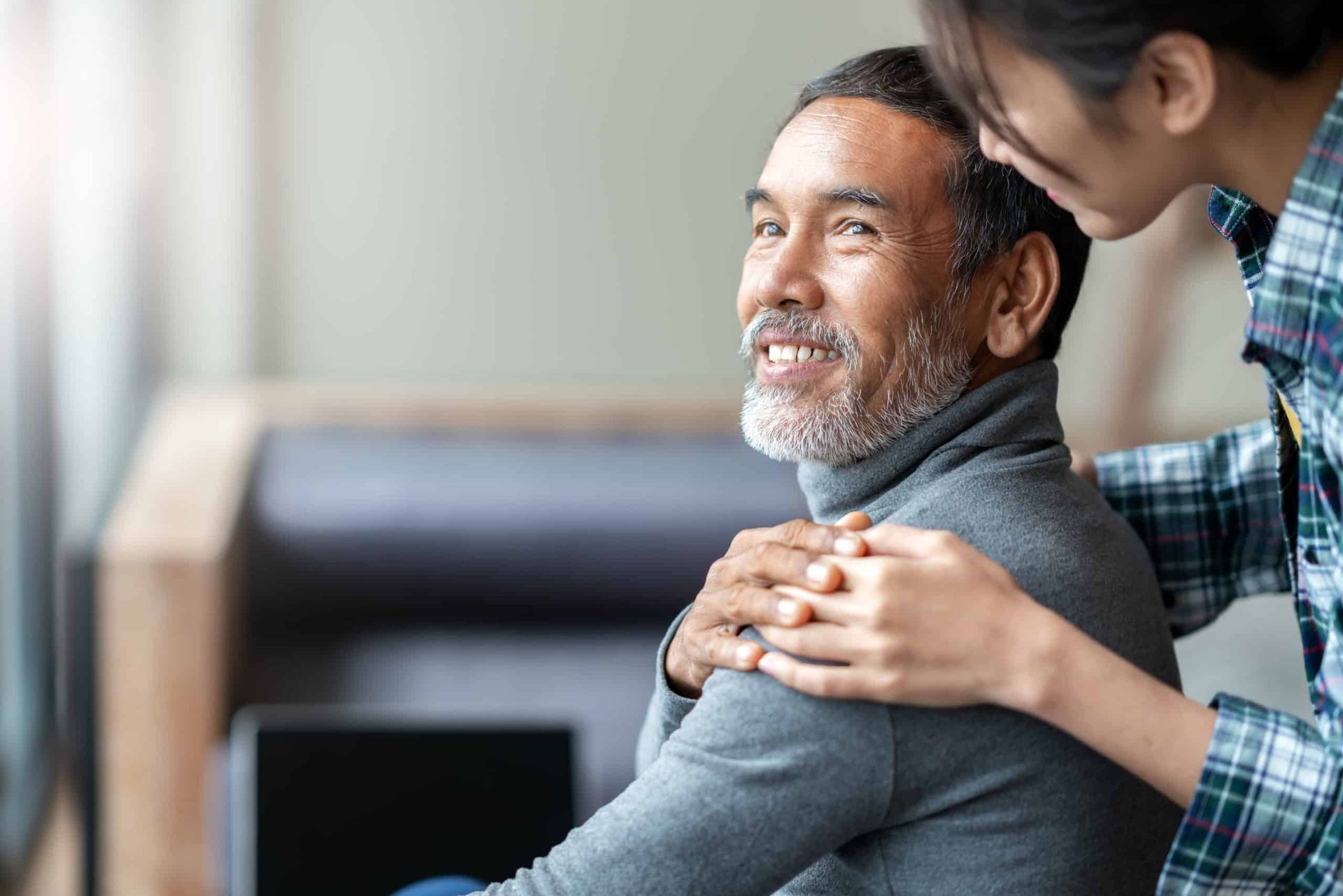 Man with graying hair smiles as a young woman puts her hands on his shoulders, indoors.