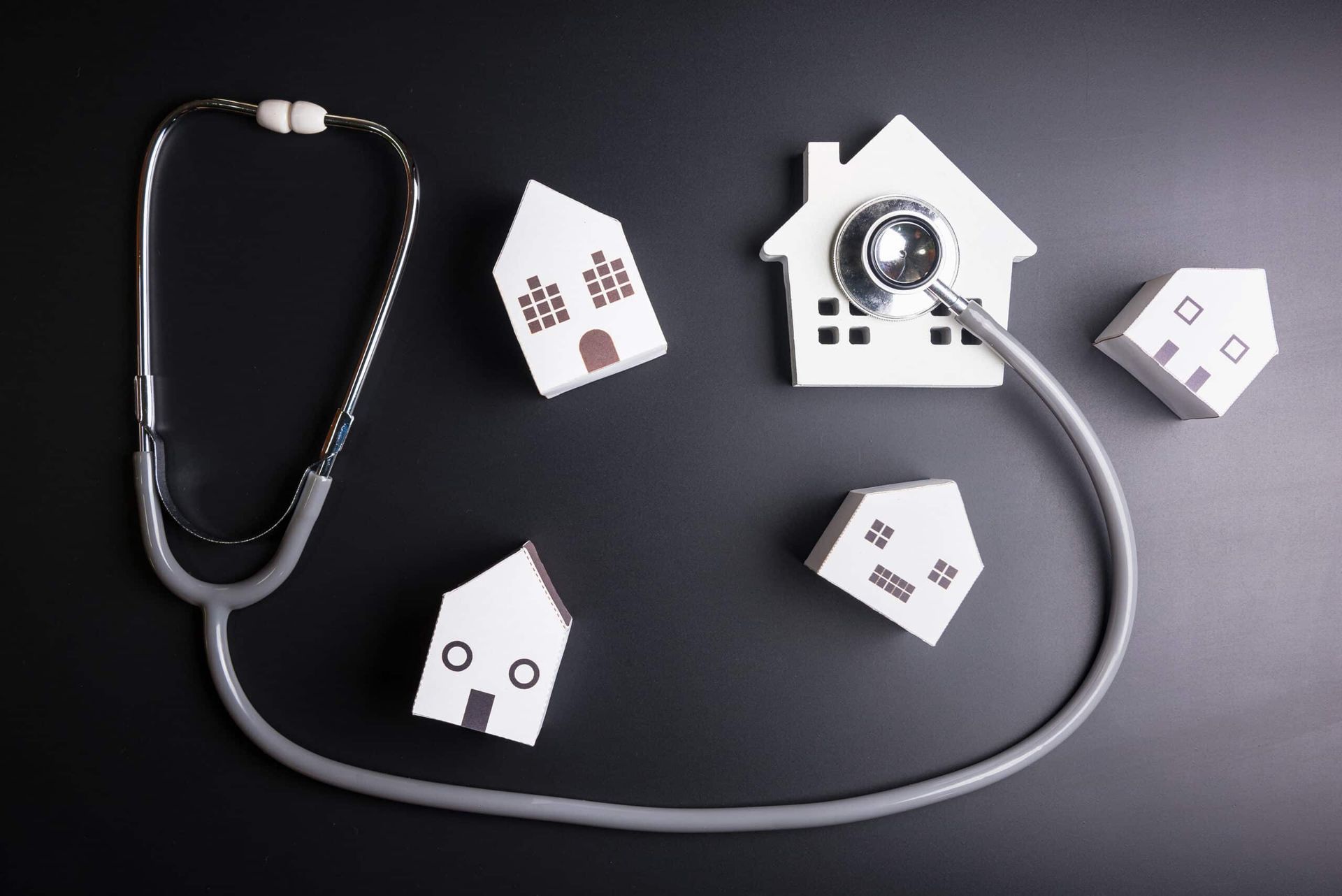 A stethoscope examines small house models on a dark surface, suggesting a home health assessment.