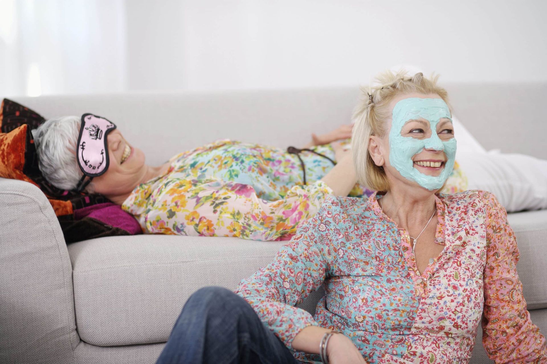 Two women relaxing on a couch; one with a face mask, smiling. The other rests with an eye mask.