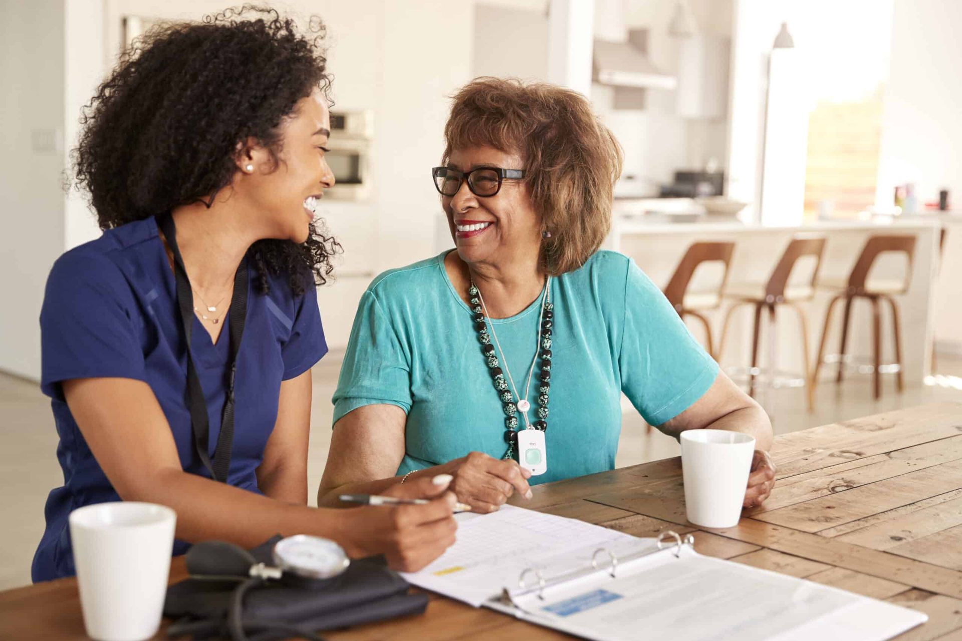 A young caregiver and elderly woman smiling at each other over paperwork at a table.