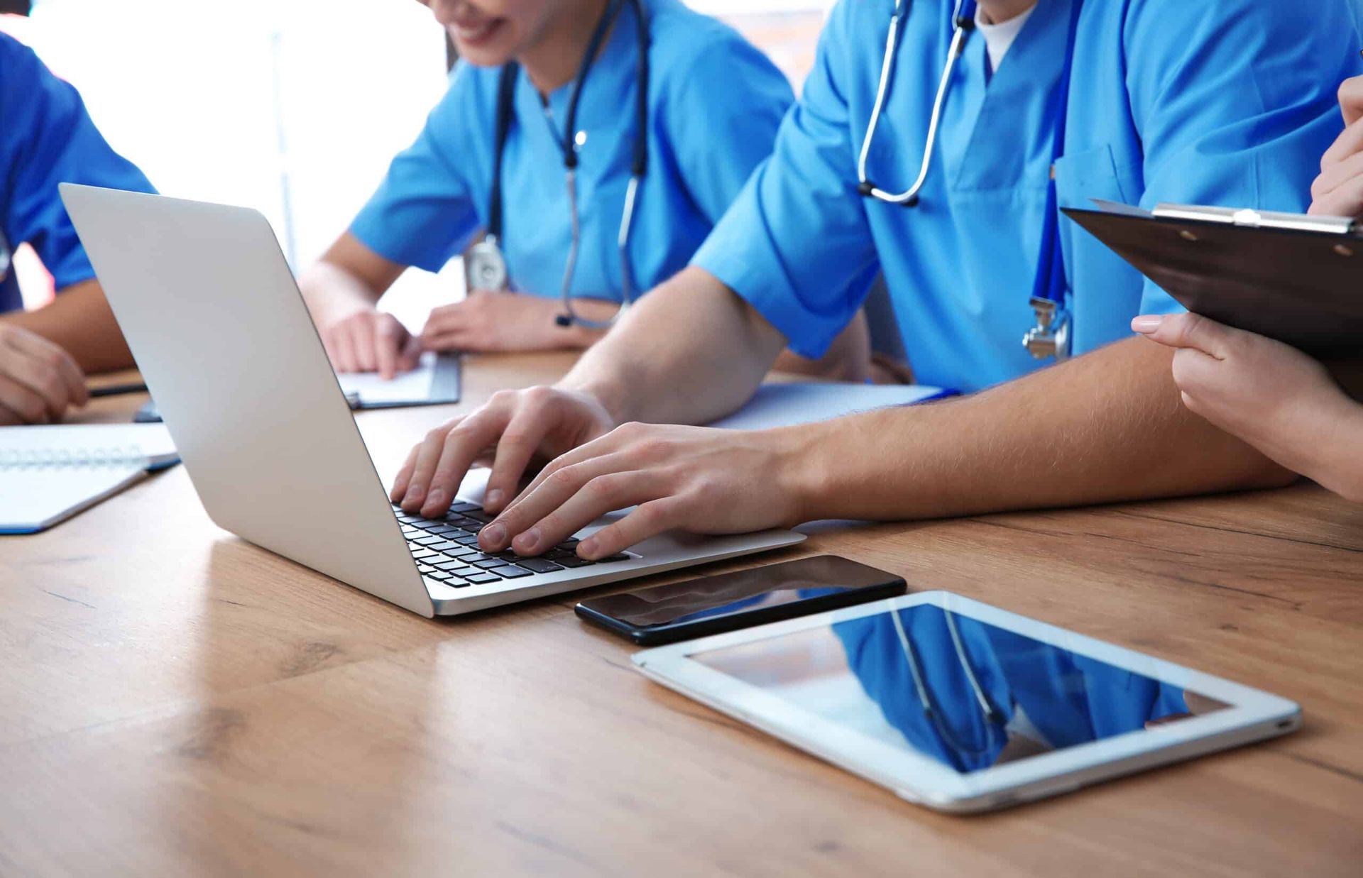 Medical team in blue scrubs using a laptop, tablet, and paperwork at a table.