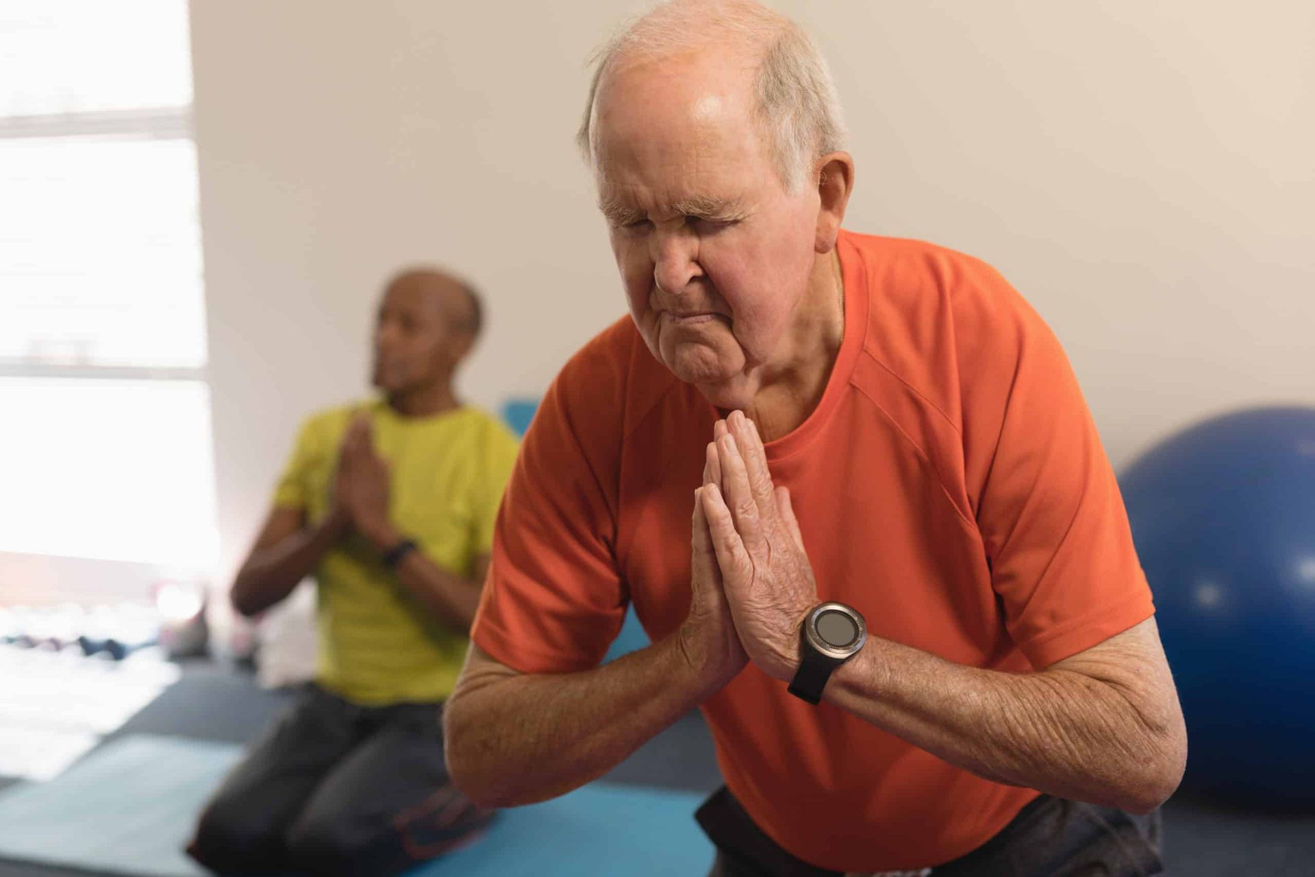 Senior man in orange shirt practicing yoga with hands together, another person in background.