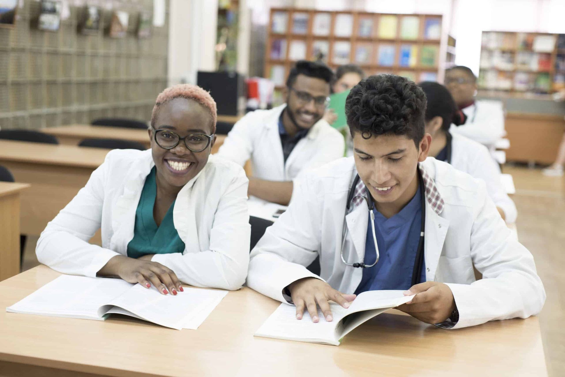Medical students studying in a classroom, smiling and looking at a book; wearing white coats.
