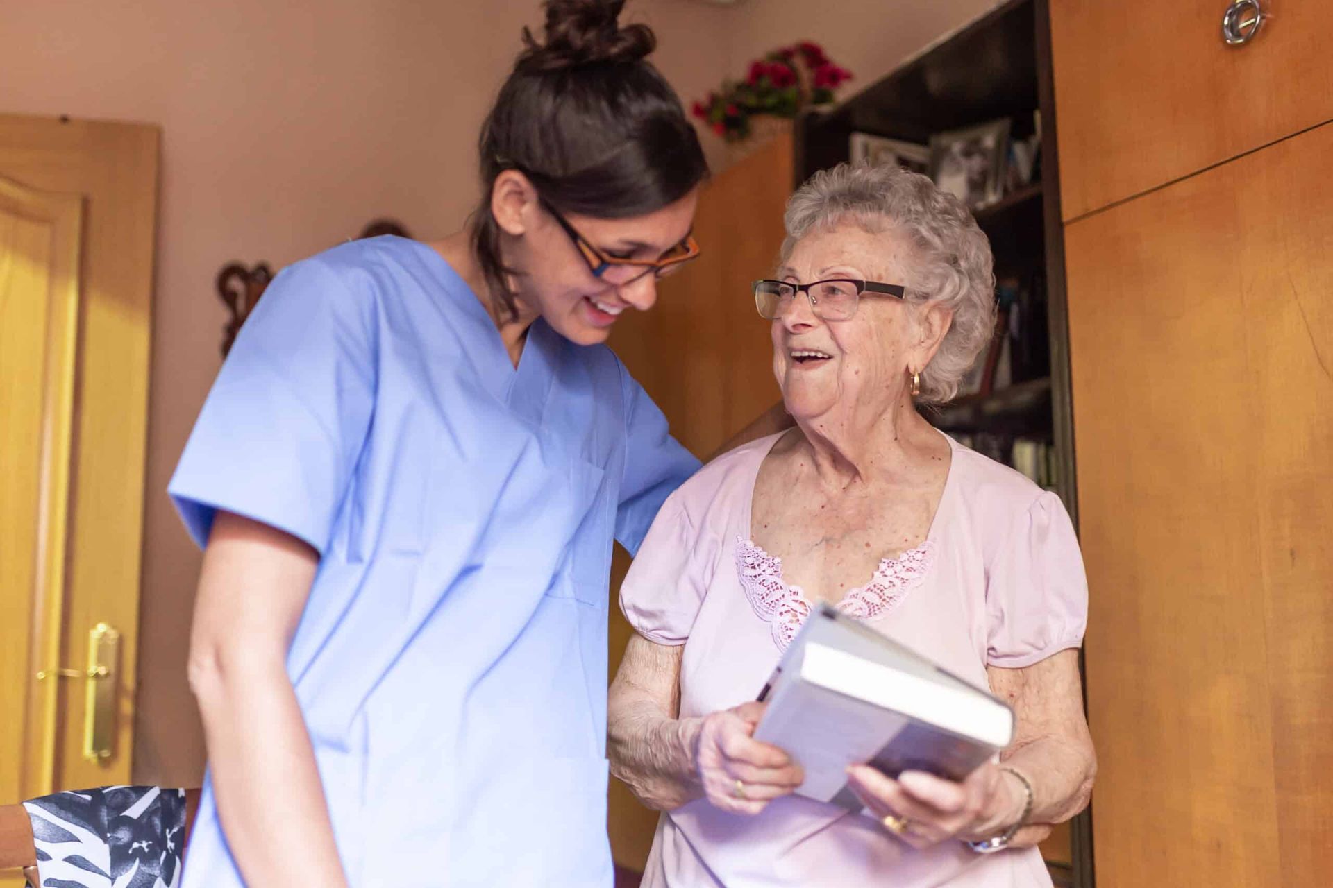 Woman in blue scrubs smiles at elderly woman holding a book indoors.