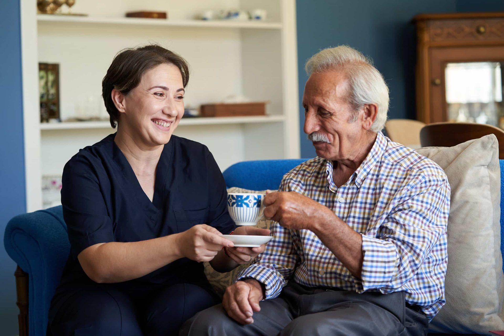 Caregiver offering a cup of tea to a smiling senior man, seated on a blue sofa in a home.