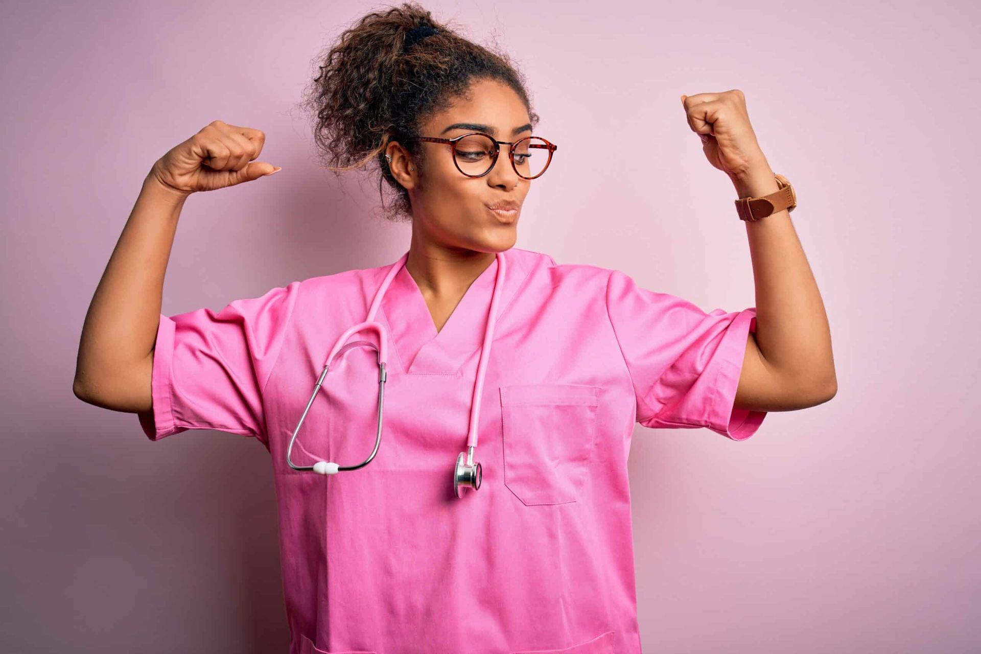 Nurse in pink scrubs flexes both arms, looking strong, pink background.