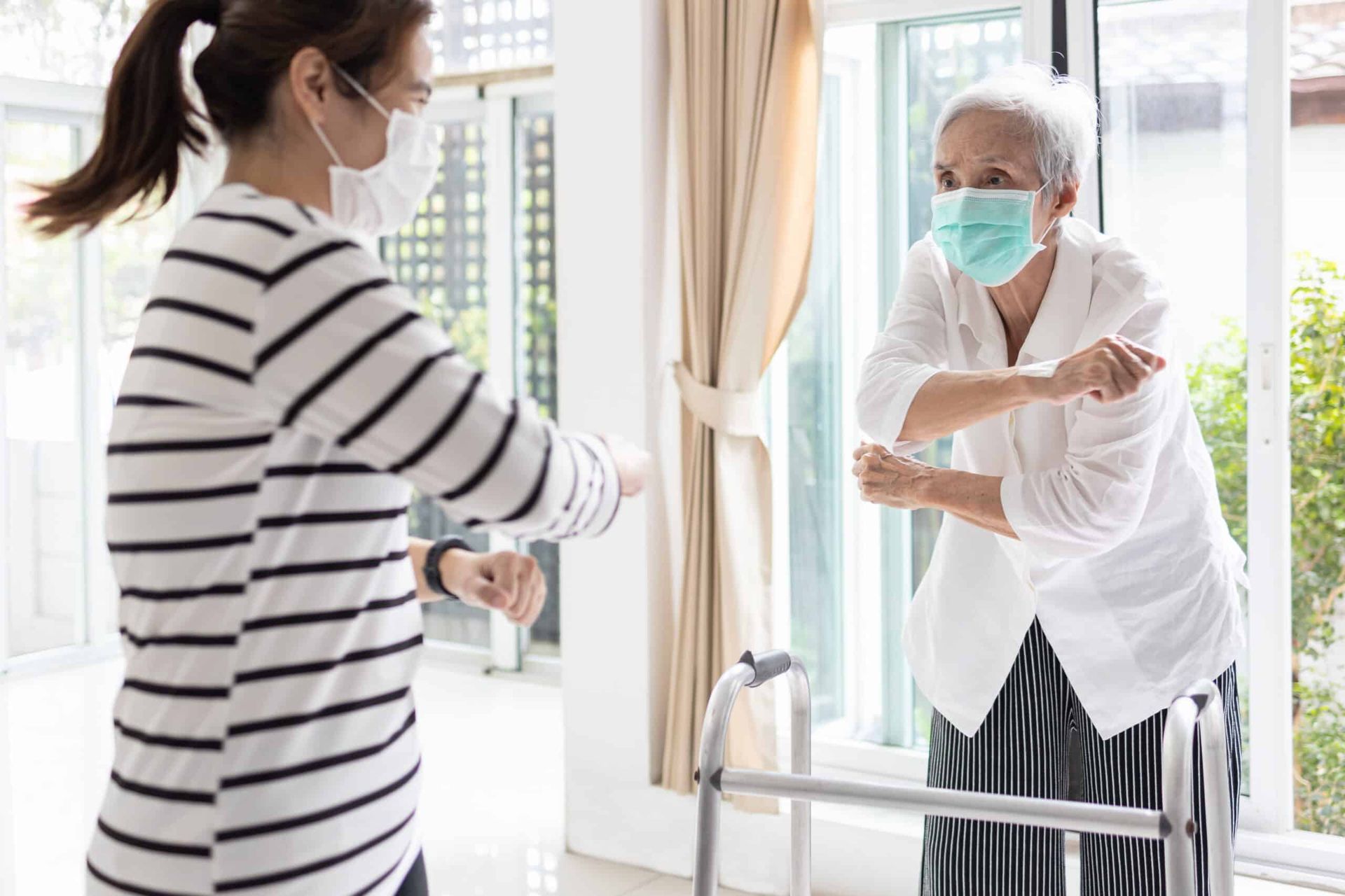 Woman and elderly woman wearing masks elbow bump inside by a window.