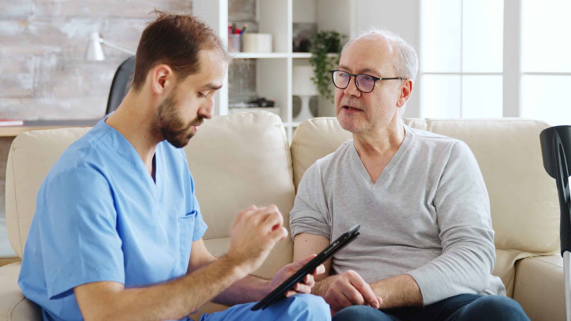 A male healthcare worker in blue scrubs reviewing a tablet with an elderly man, seated on a couch.