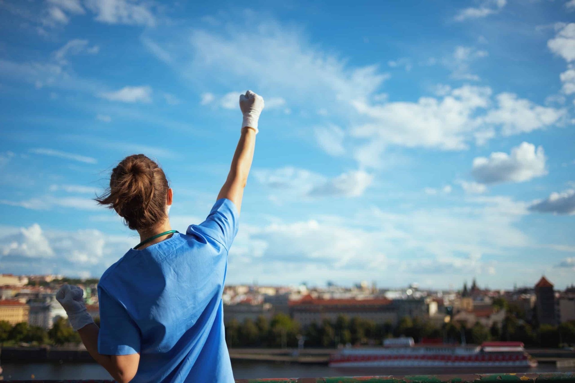Medical worker raises fist in triumph against a sunny sky and cityscape.
