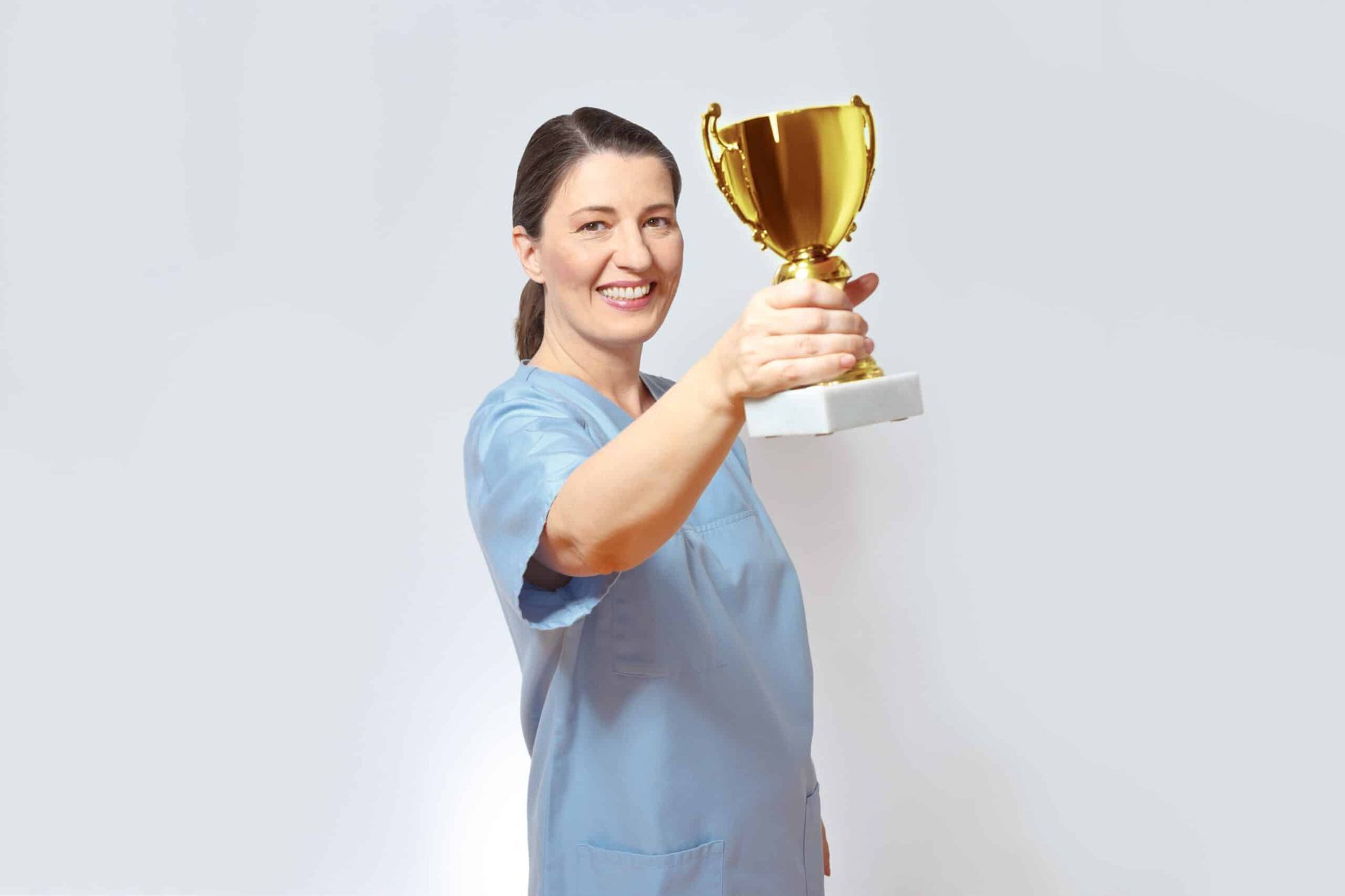 Smiling woman in blue scrubs holding up a gold trophy against a white background.