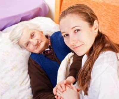 Young woman holding hands with elderly woman in bed; both smiling.