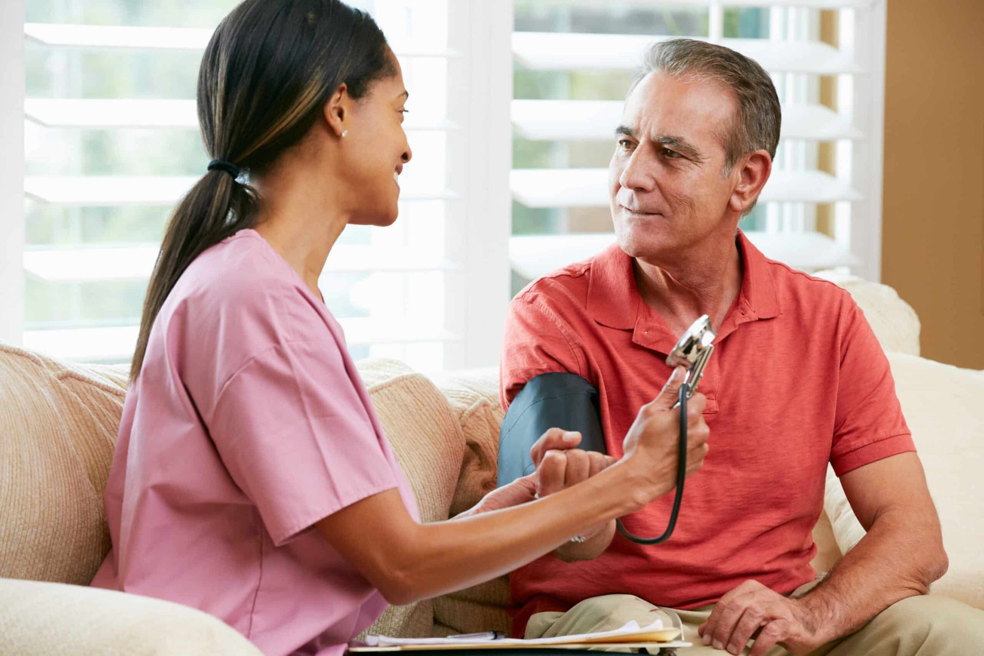 Nurse taking a man's blood pressure at home. Nurse in pink scrubs smiles, man in orange shirt looks on.