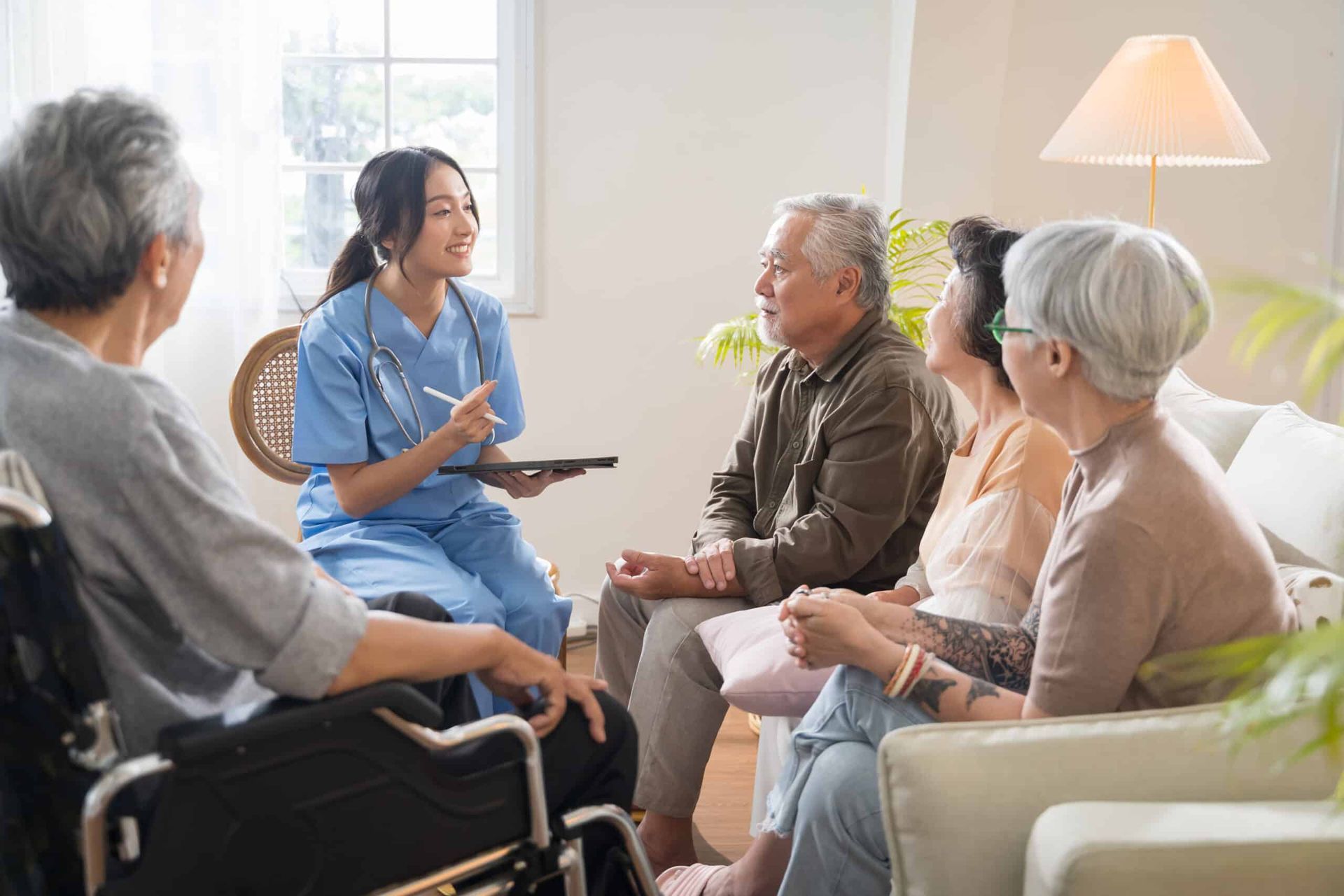 Nurse in scrubs talking with a group of seniors in a living room; one uses a wheelchair.